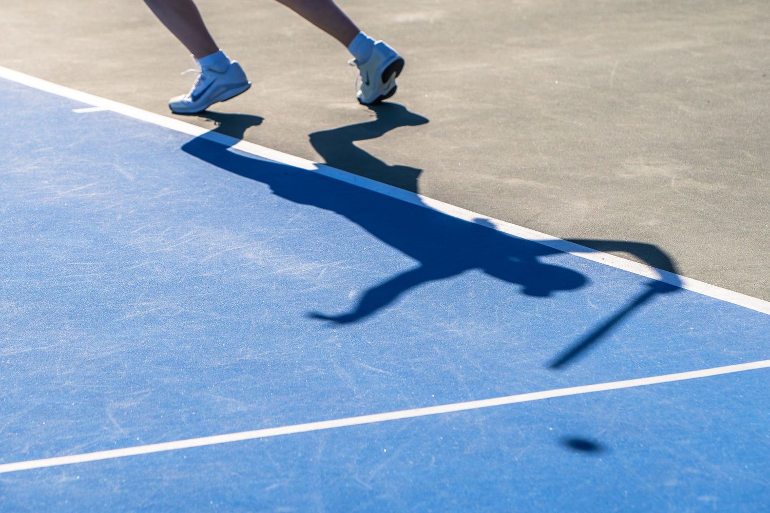 Shadow of a tennis player and racket cast on a bright blue outdoor tennis court surface.