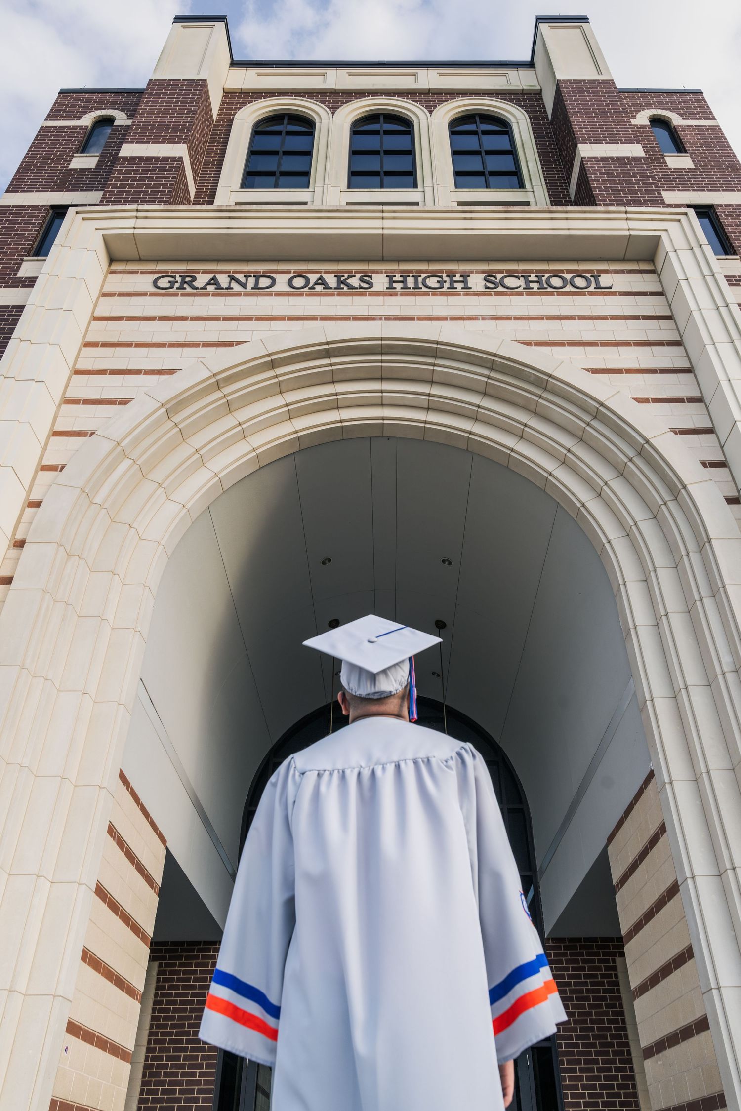 Graduate in white cap and gown stands before Grand Oaks High School entrance archway.