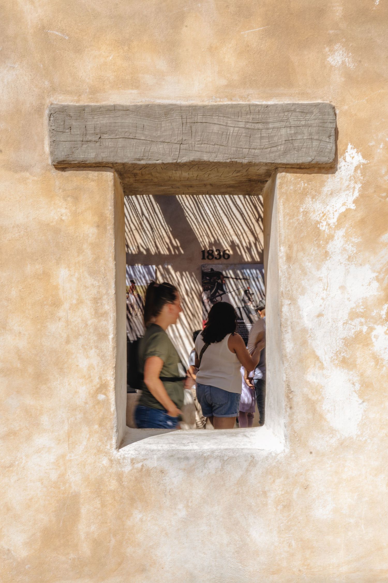 Tourists visible through a rustic adobe window opening with wooden beam lintel at a historic 1836 southwestern building.
