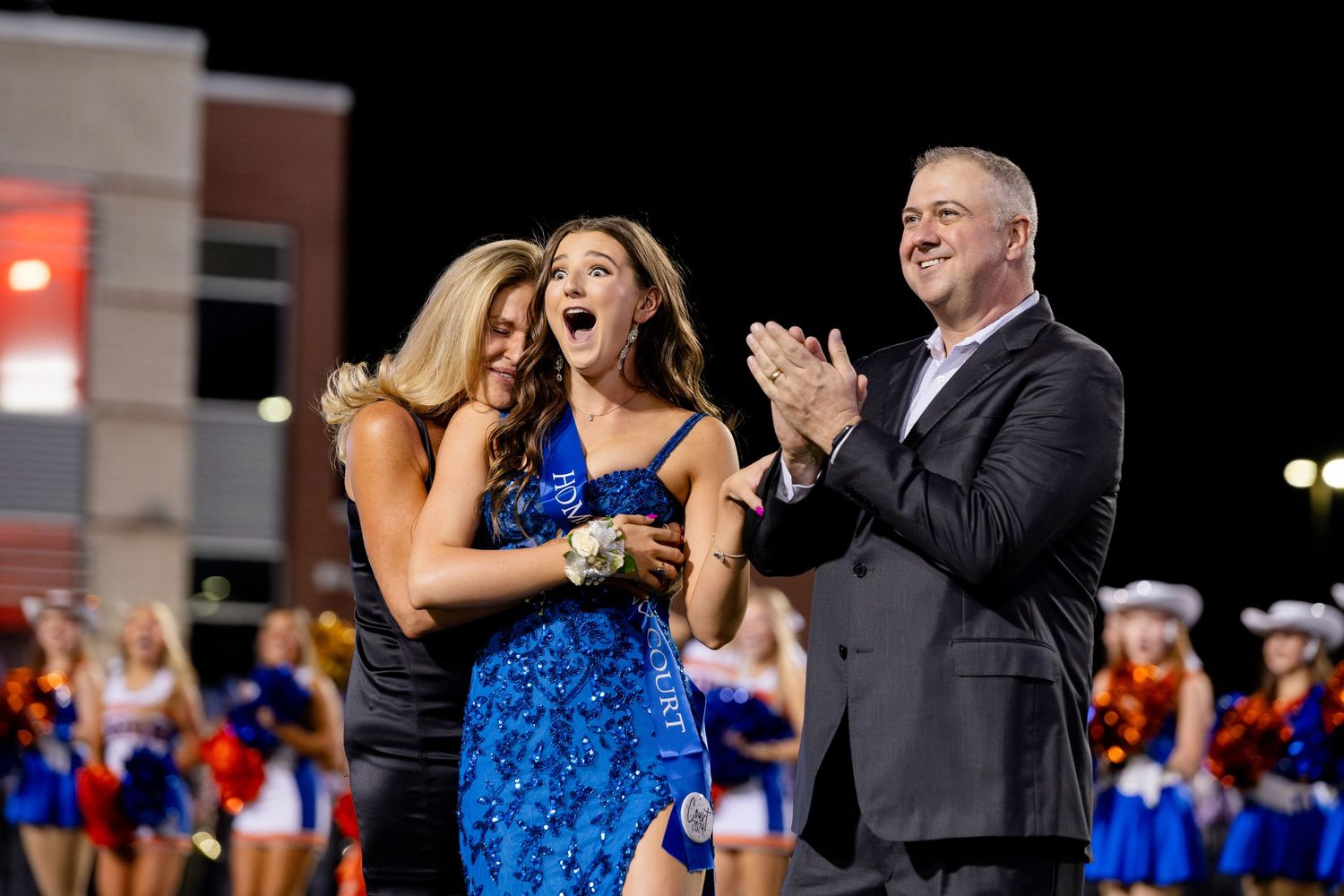 A surprised young woman in a blue sequined dress reacts joyfully at a homecoming ceremony on a football field at night.