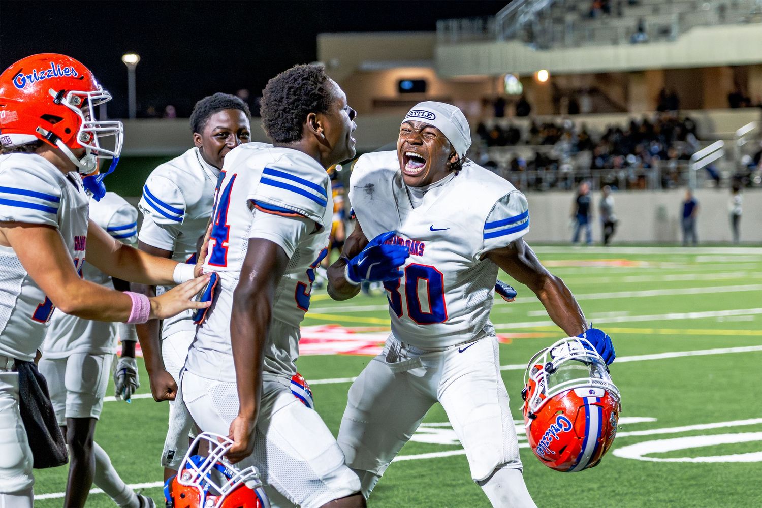 Football players in blue and white uniforms celebrate excitedly on the field during a nighttime game.