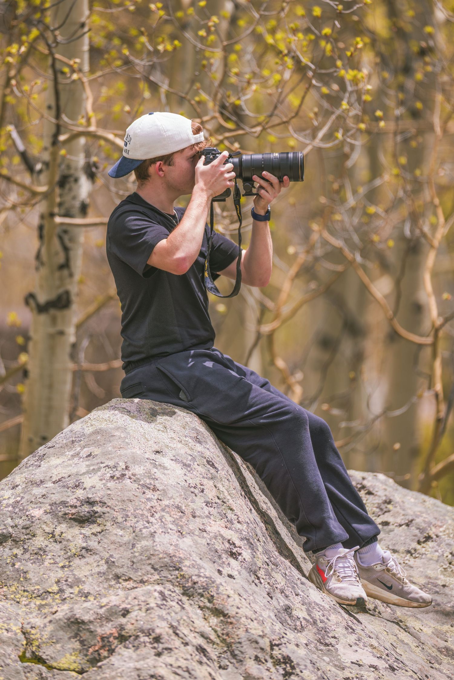 Photographer in a baseball cap sits on a boulder, shooting with a DSLR camera in an autumn forest.