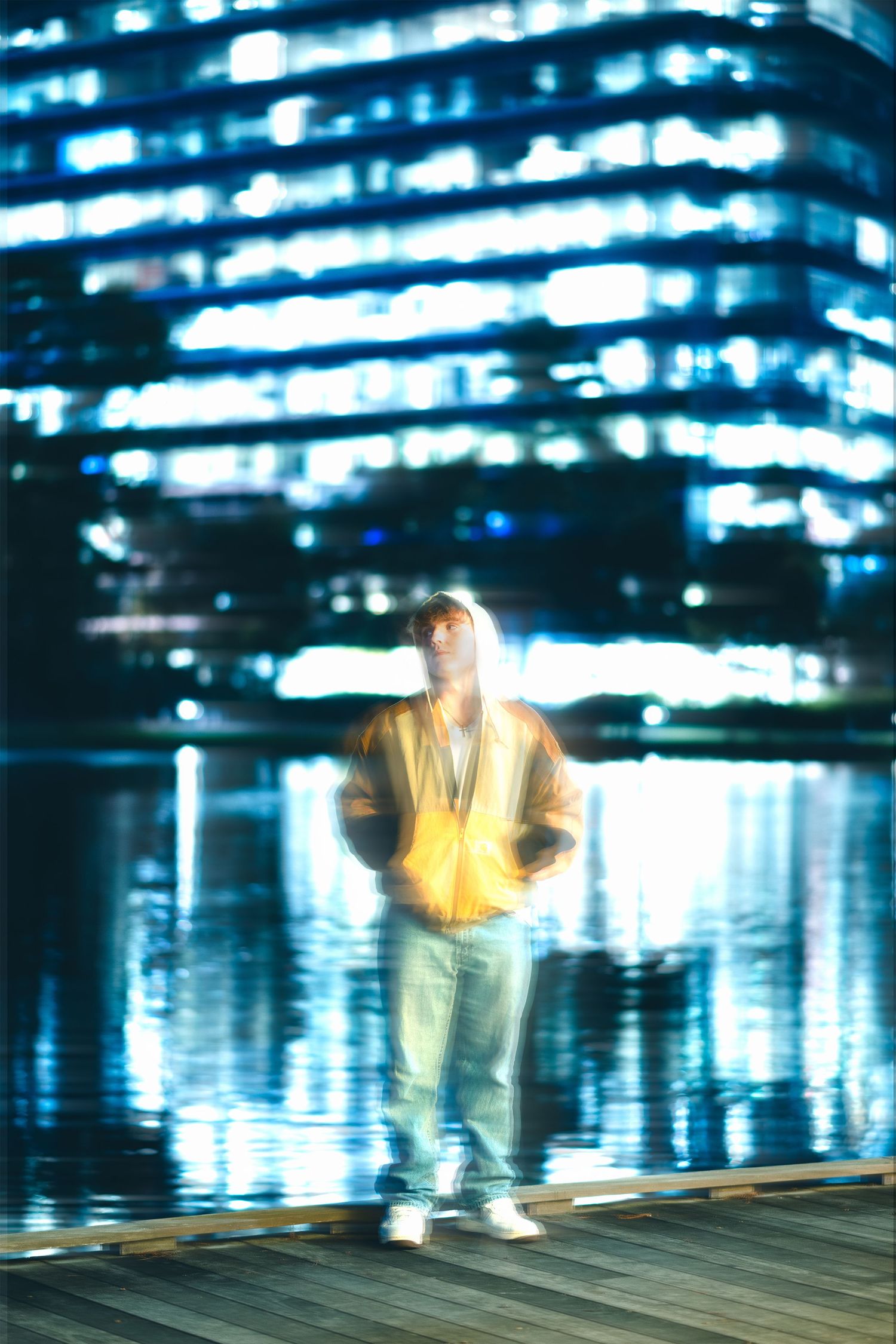 A person in a yellow jacket stands near a reflective waterfront with a glowing blue skyscraper in the background at night.