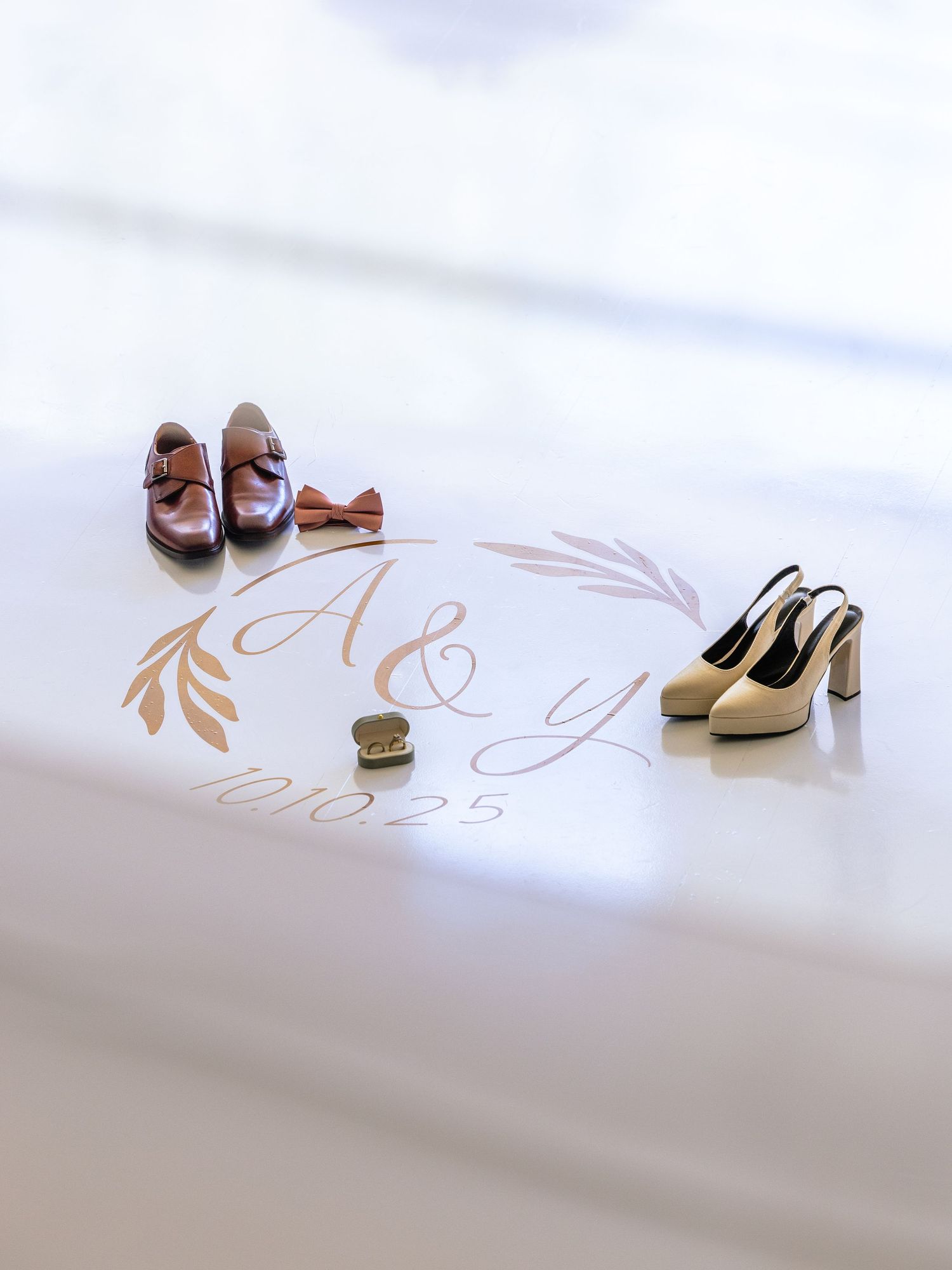 Wedding flat lay with monogram A&Y 10.10.25, brown dress shoes, bow tie, gold rings, and beige heels on white surface.