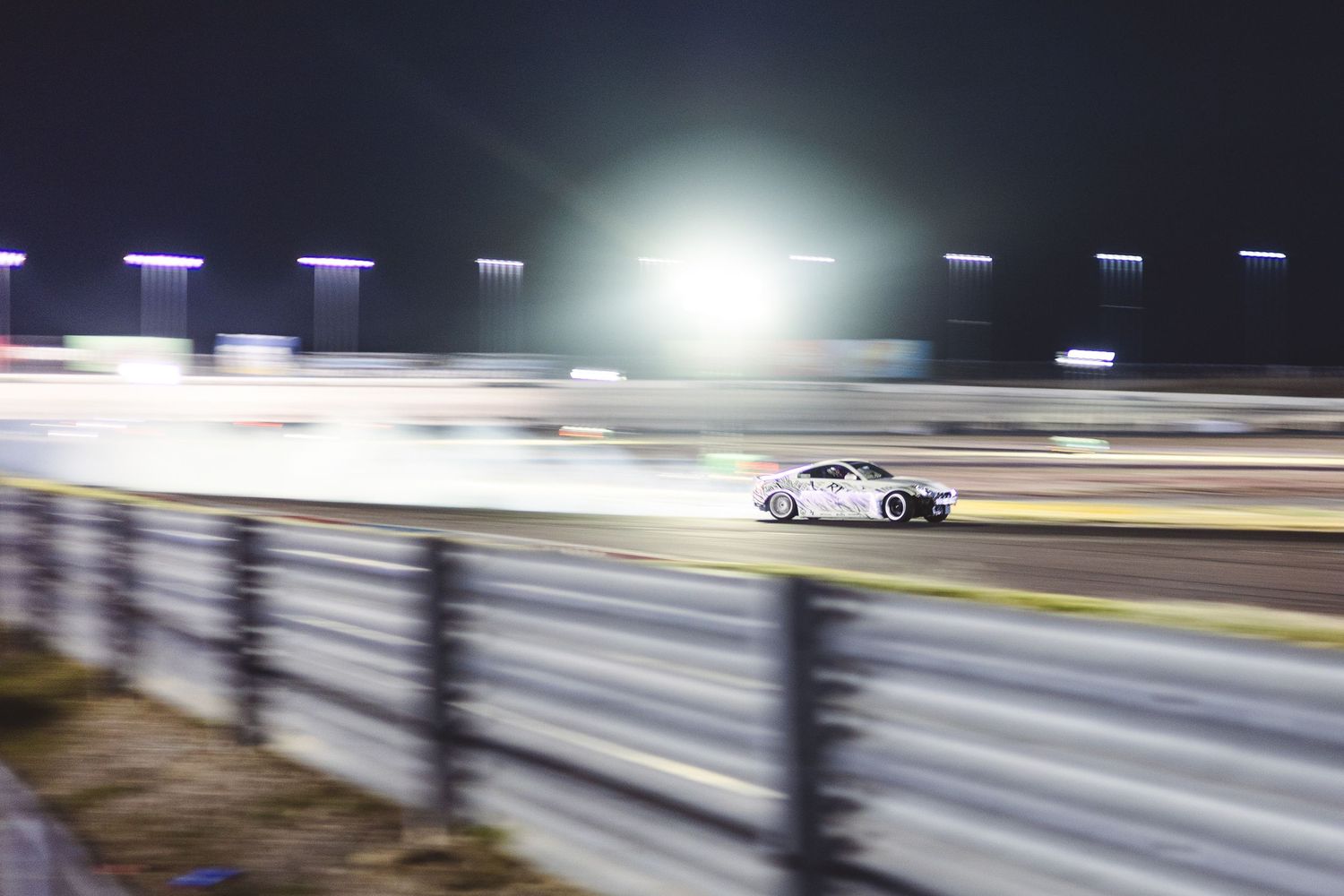 A white race car drifts on a night circuit with bright stadium lights illuminating the track.