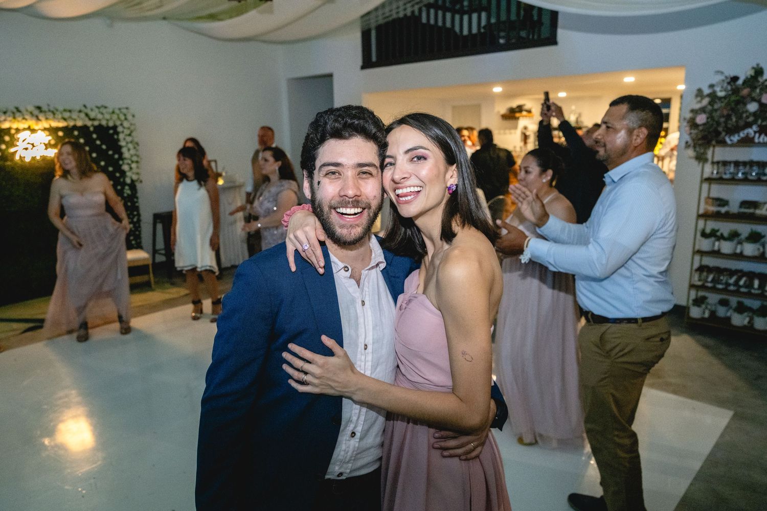 Couple smiling and embracing on a wedding dance floor with guests celebrating in the background.