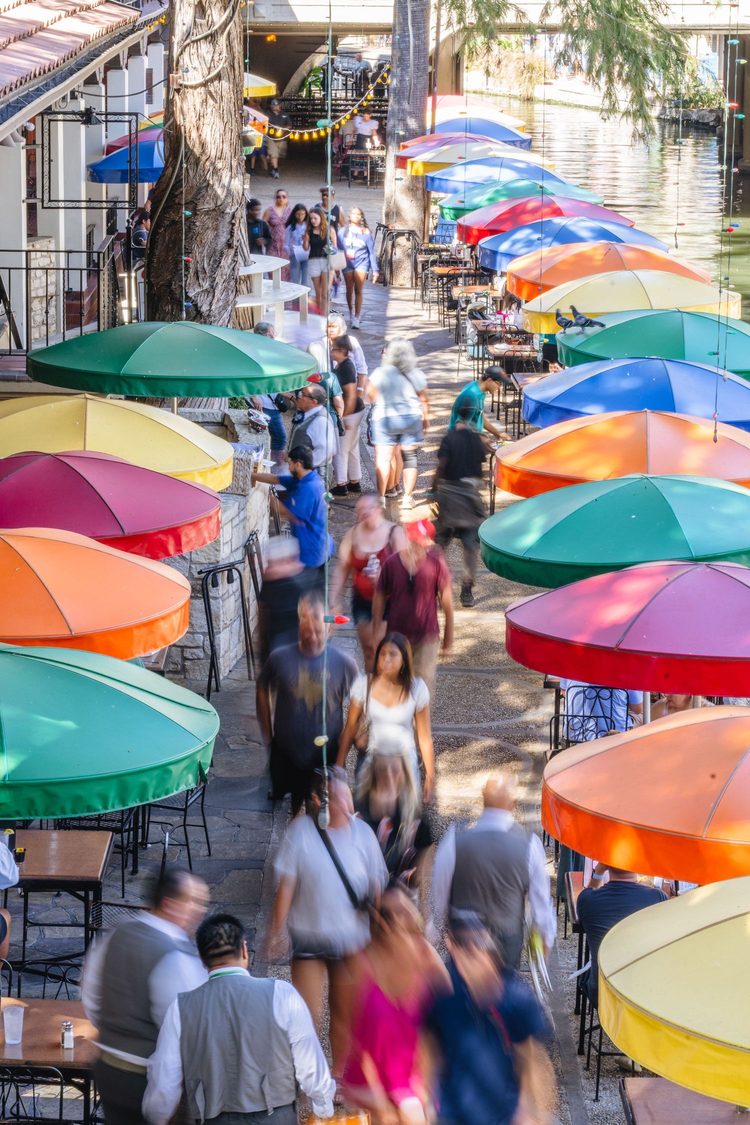 Aerial view of crowded San Antonio River Walk lined with colorful umbrellas and outdoor dining.