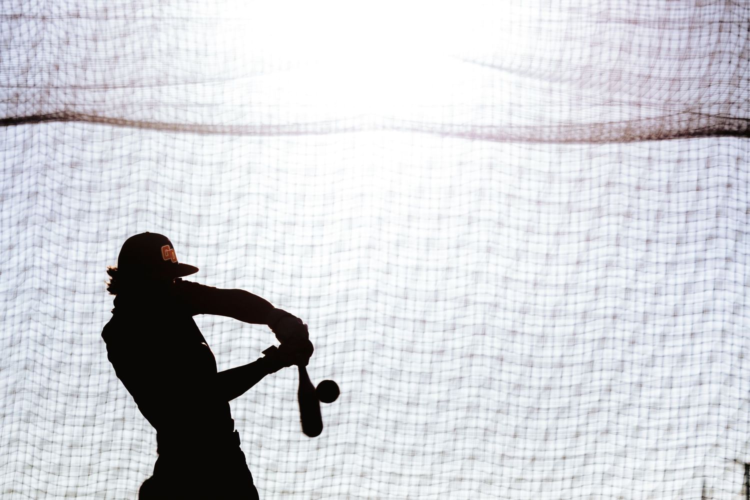 Silhouette of a baseball batter swinging against a bright backlit batting cage net.