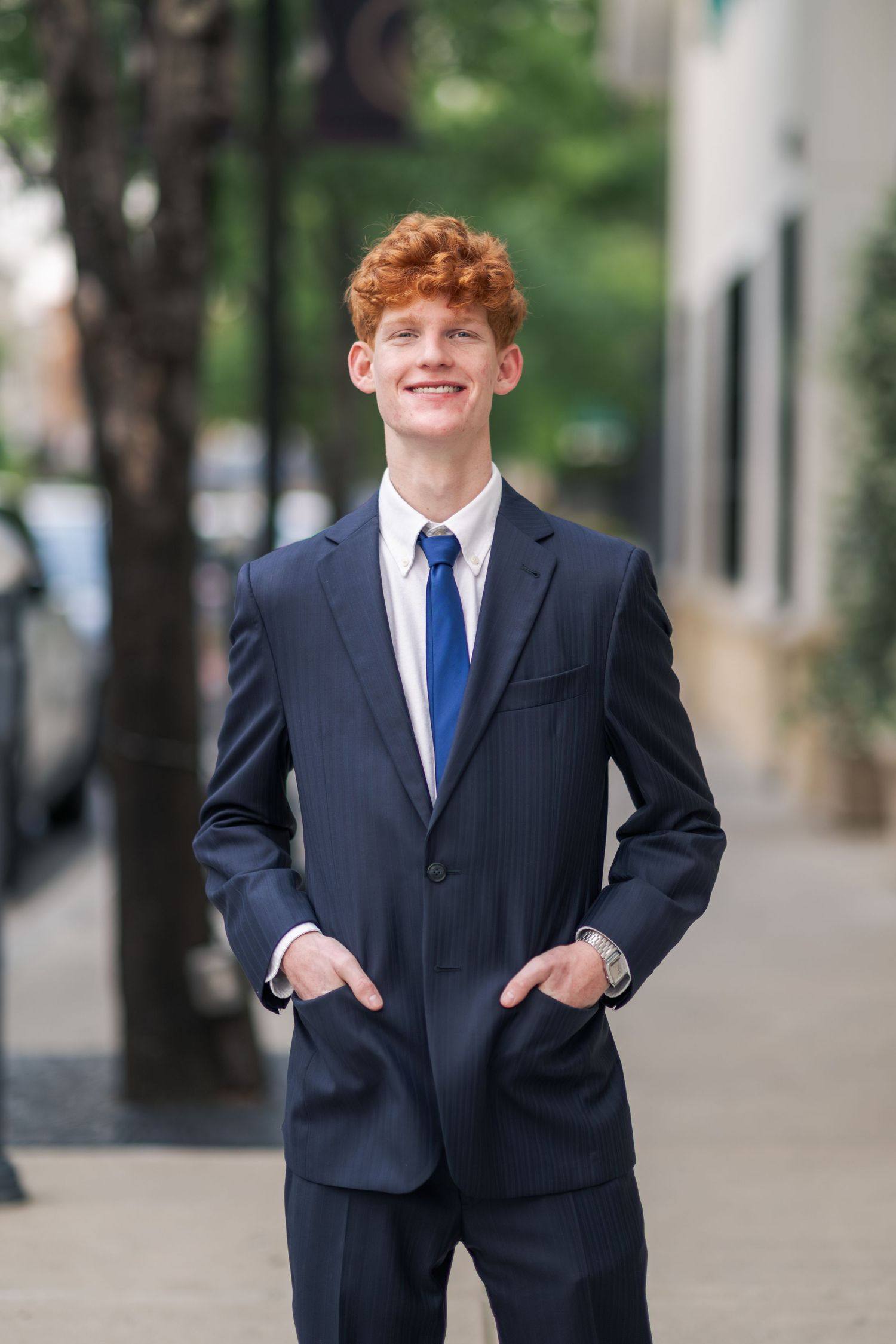 Smiling young man with red hair in navy suit and blue tie poses on city sidewalk.