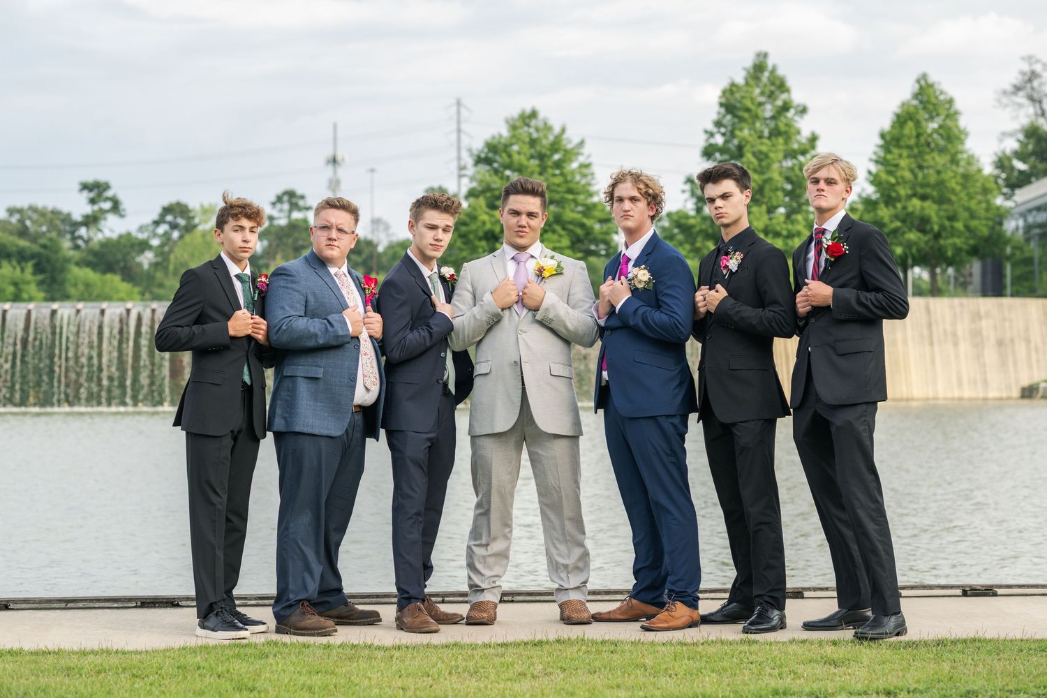 Six young men in suits and tuxedos posing together at prom near waterfront backdrop.
