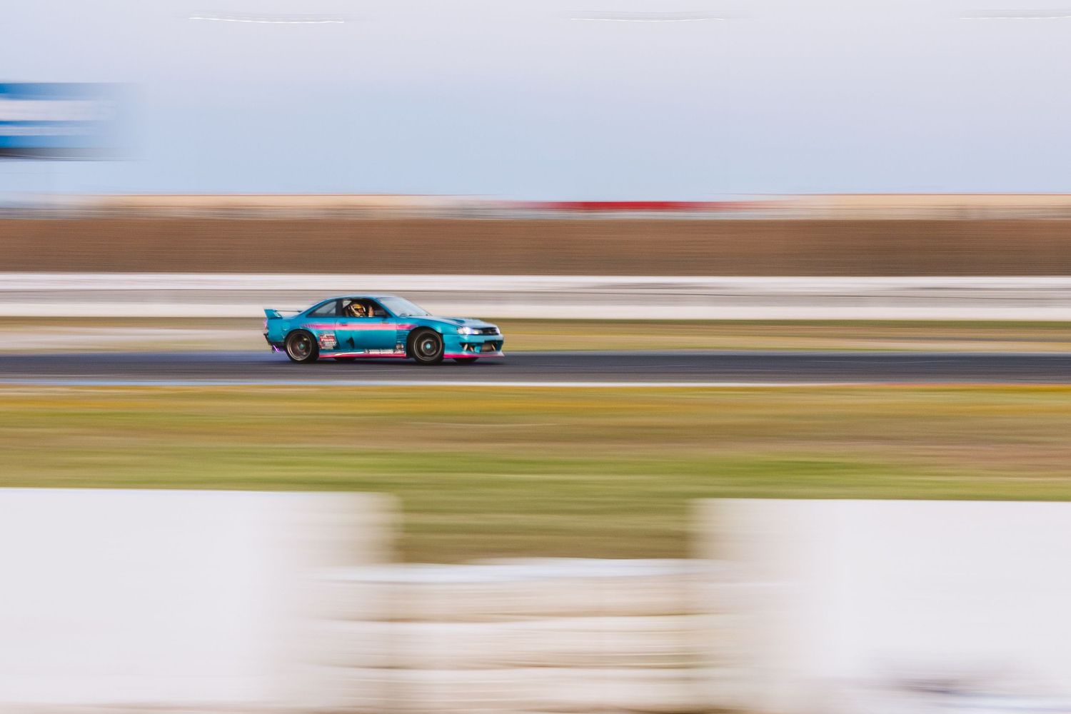 A light blue race car captured with panning technique speeding along a racing circuit during daytime.