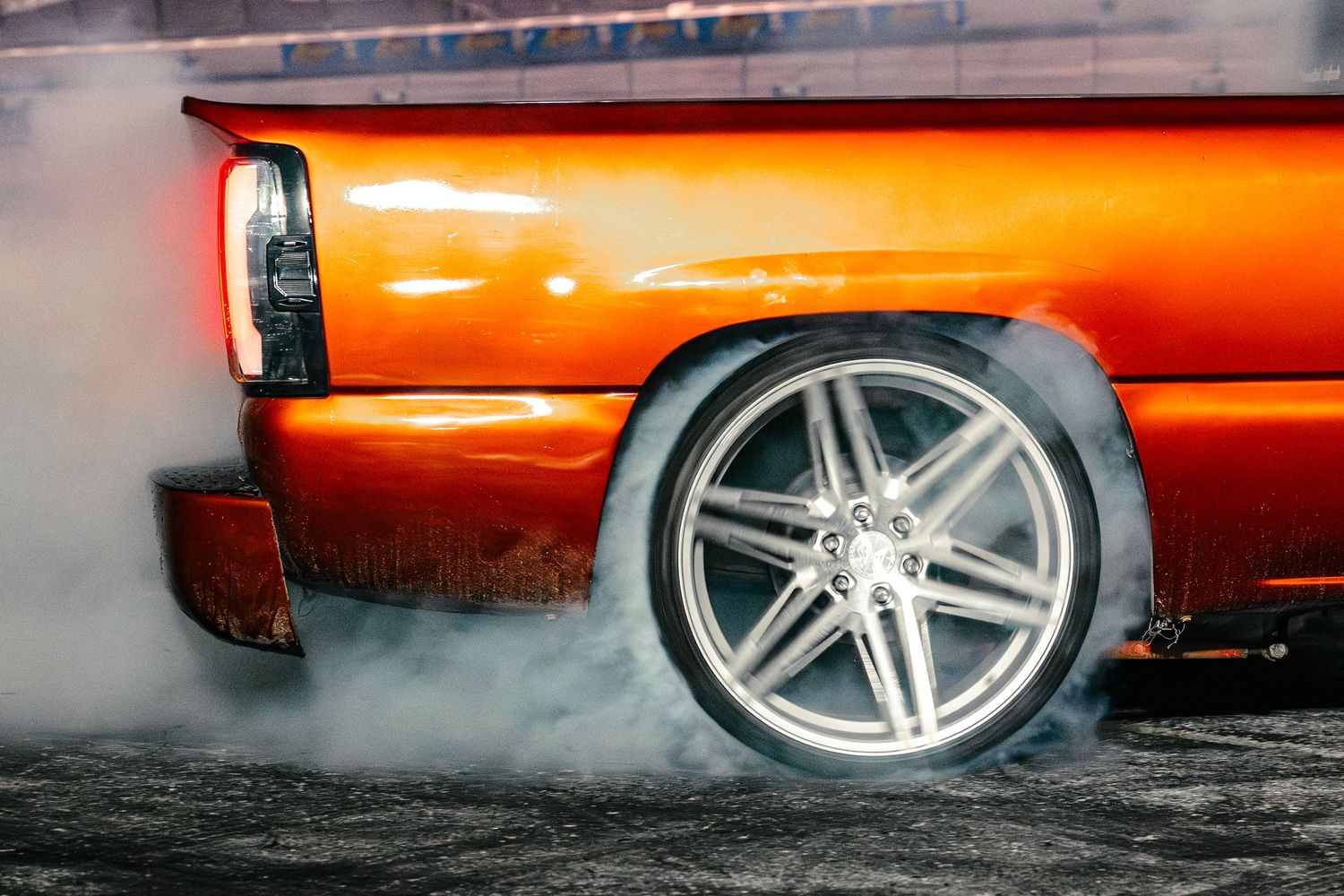 Close-up of a chrome wheel on a custom orange truck performing a burnout with thick white tire smoke.