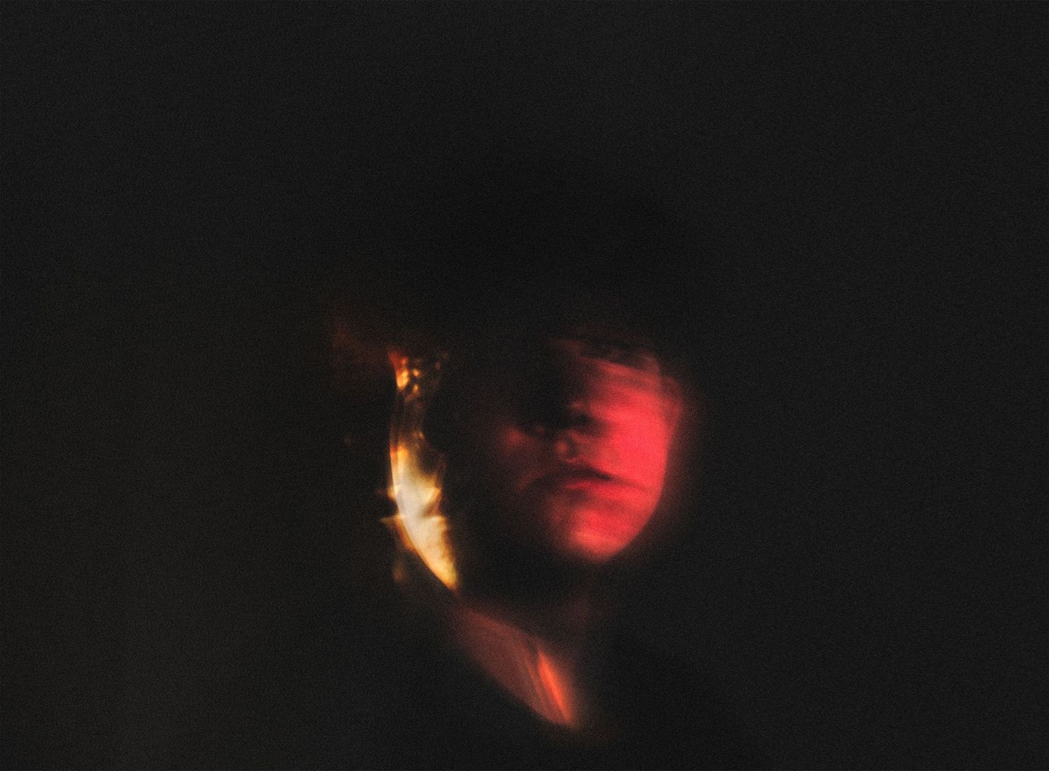 A dark moody portrait with motion blur showing a persons face illuminated by warm red and yellow tones.