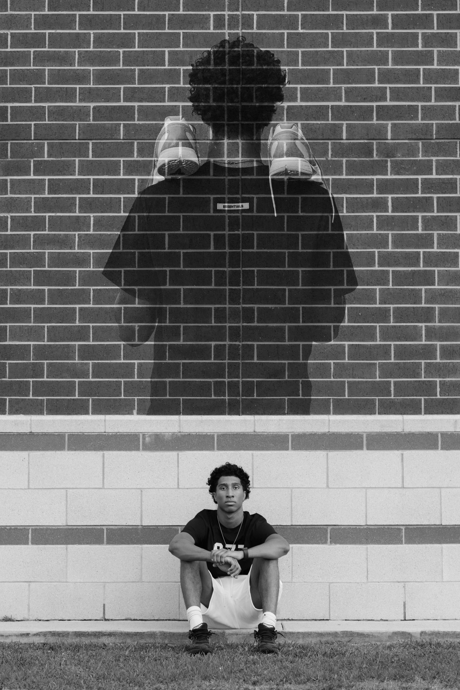 Black and white photo of a young man sitting against a brick wall with his shadow looming large behind him.
