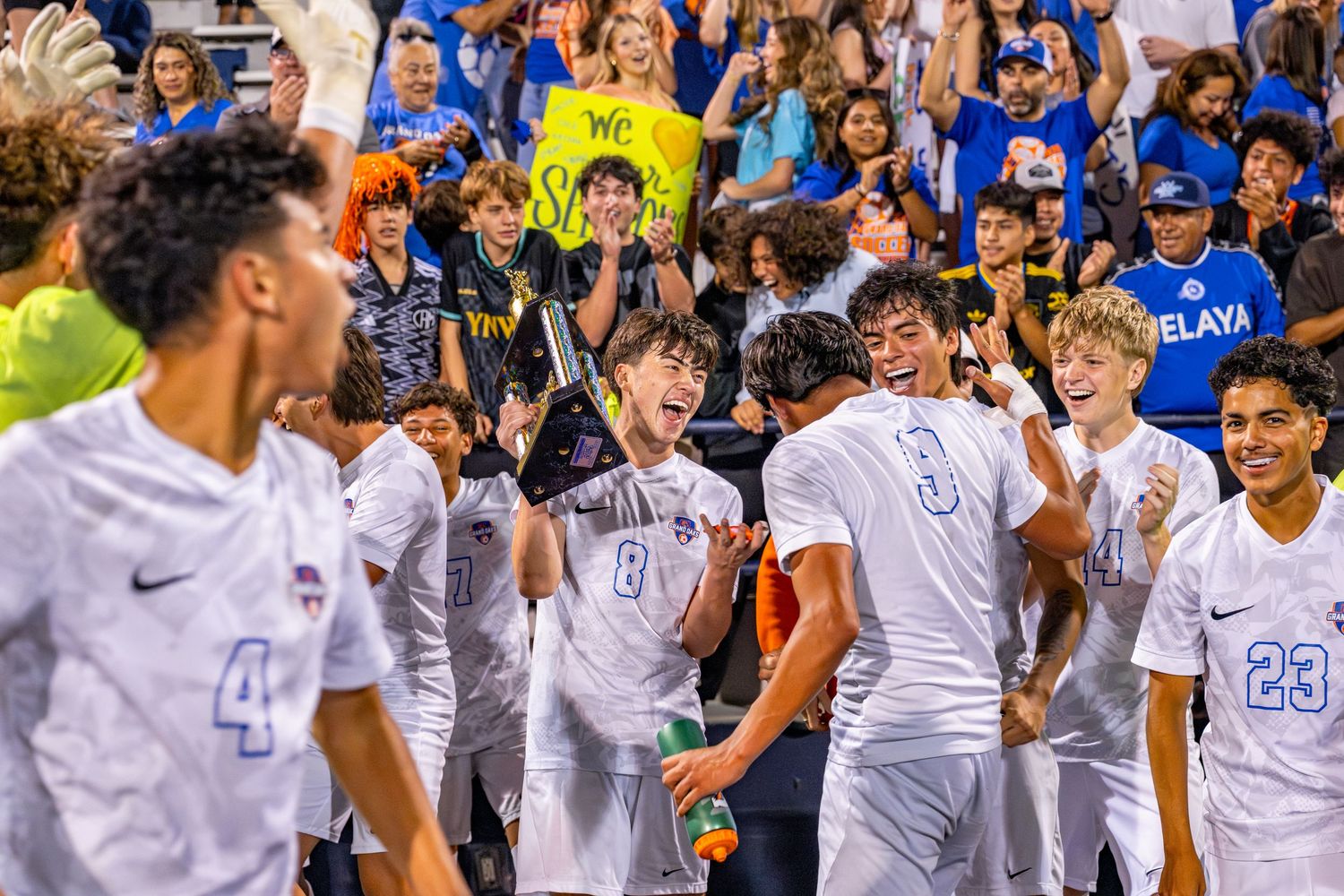 Soccer players celebrate victory with cheering fans holding signs in packed stadium stands.