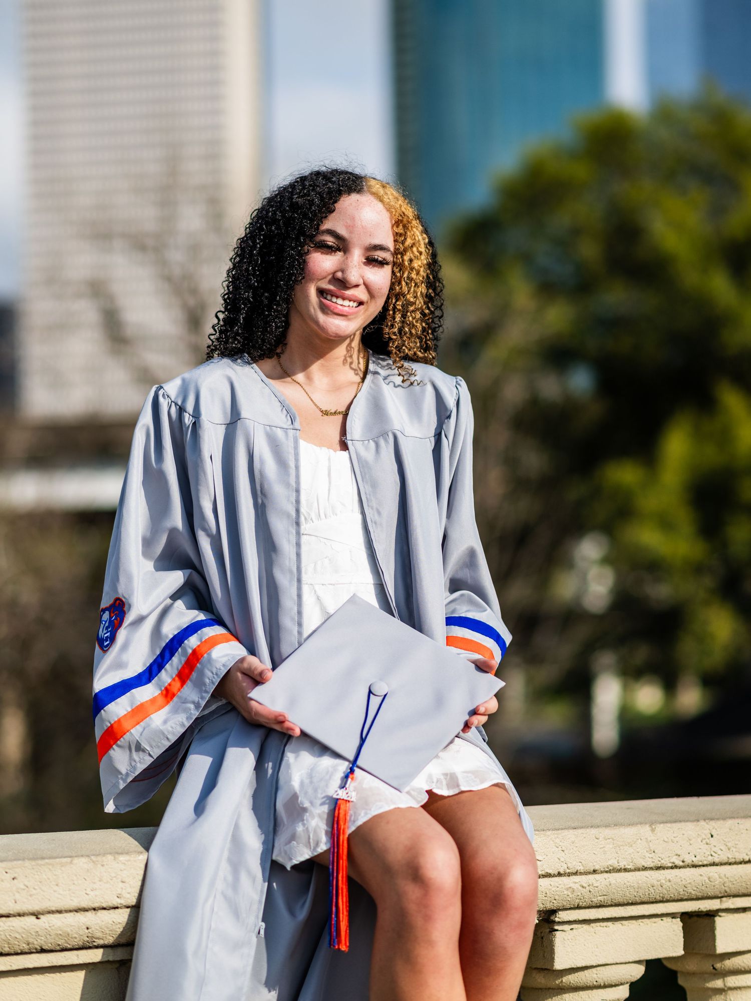 Smiling young woman in gray graduation gown holding cap outdoors in urban setting.