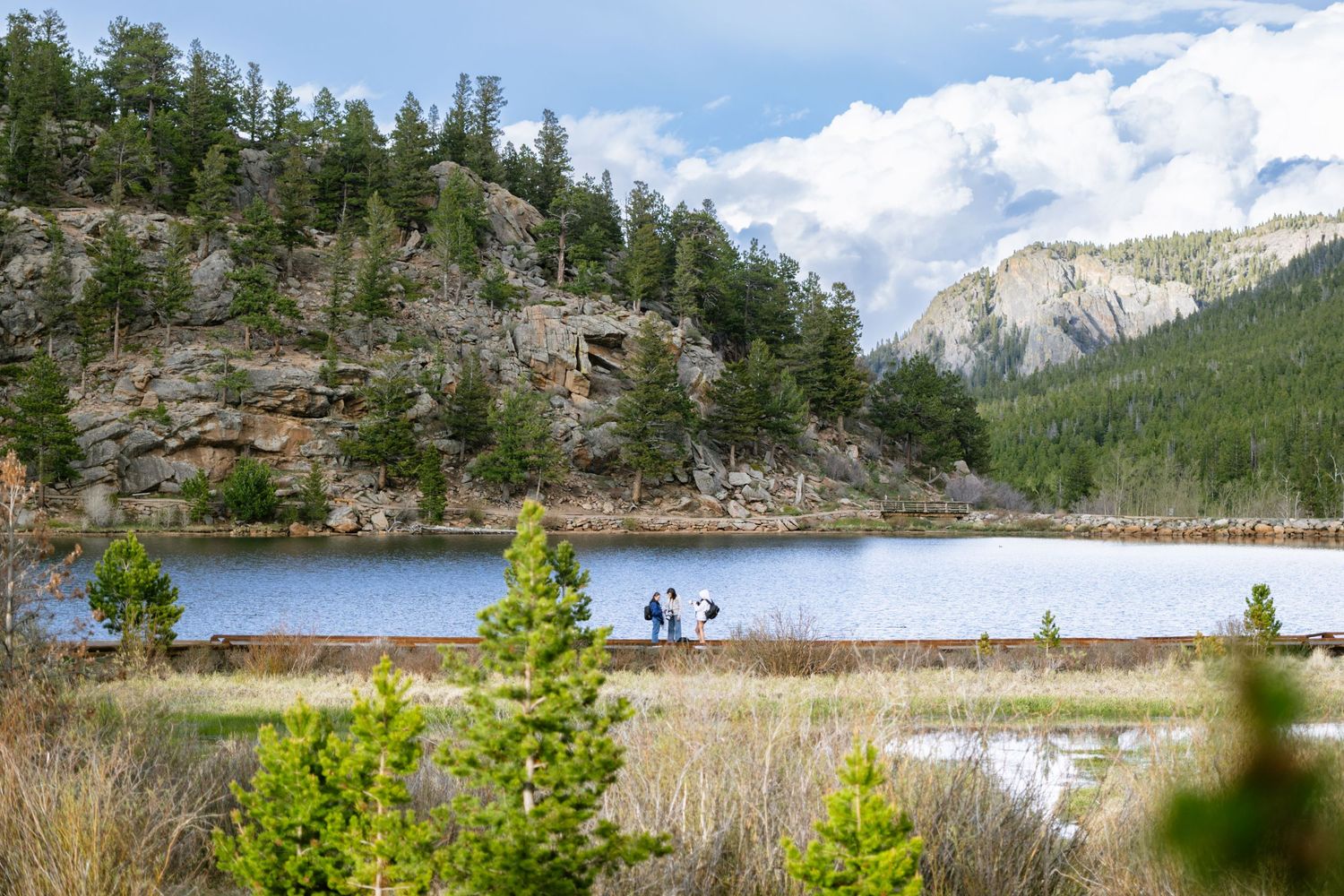 Scenic mountain lake surrounded by pine trees with two visitors on the shoreline path.
