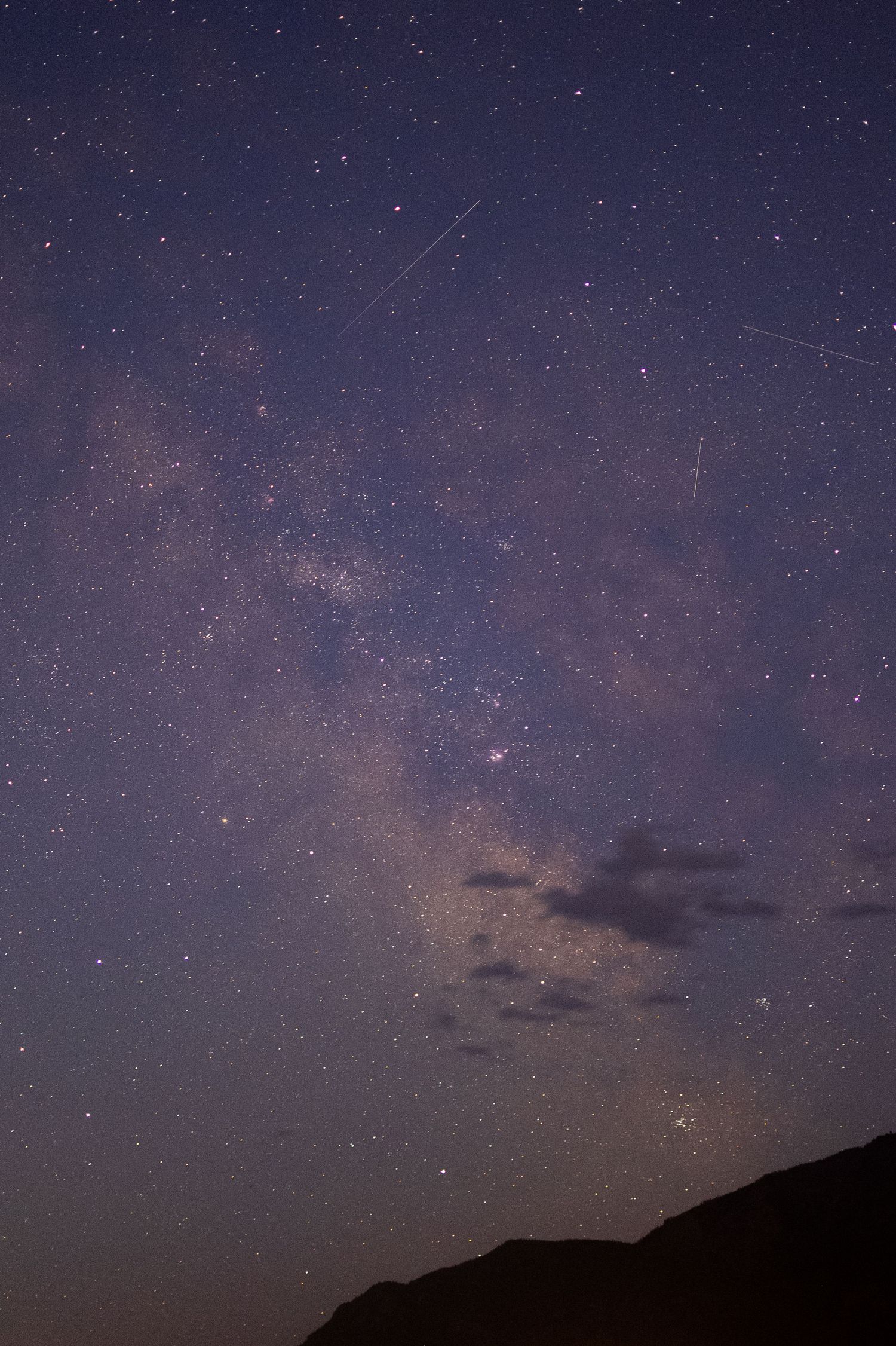 Breathtaking Milky Way galaxy with shooting stars visible over a dark mountain silhouette.