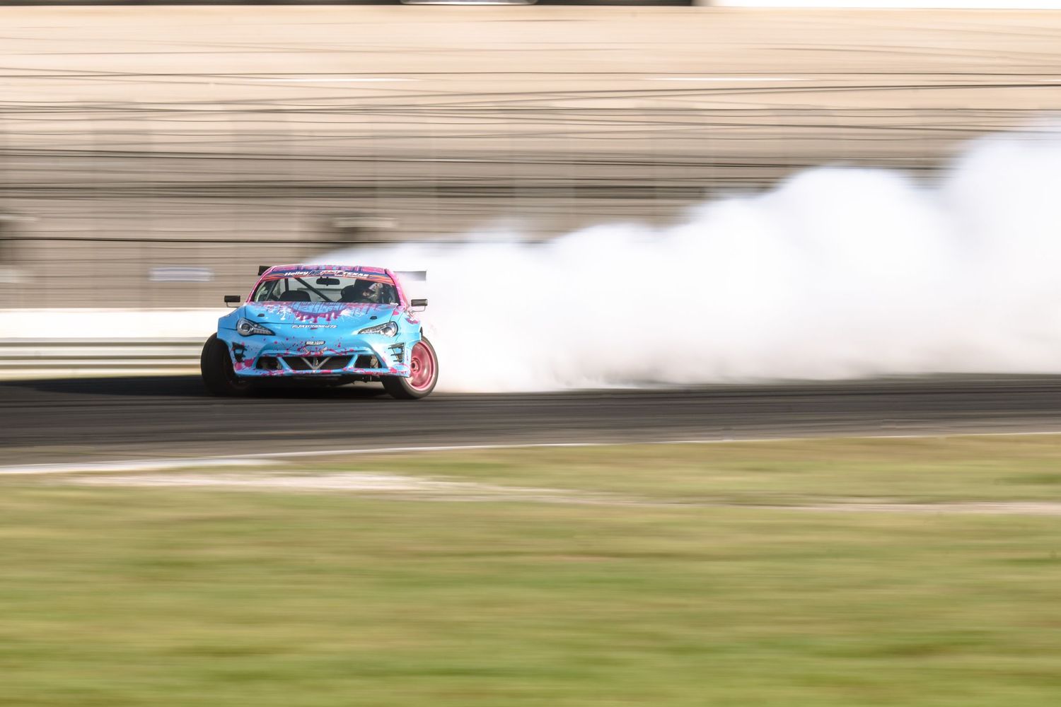 Blue drift car sliding through a corner with heavy tire smoke on a sunny racing circuit.