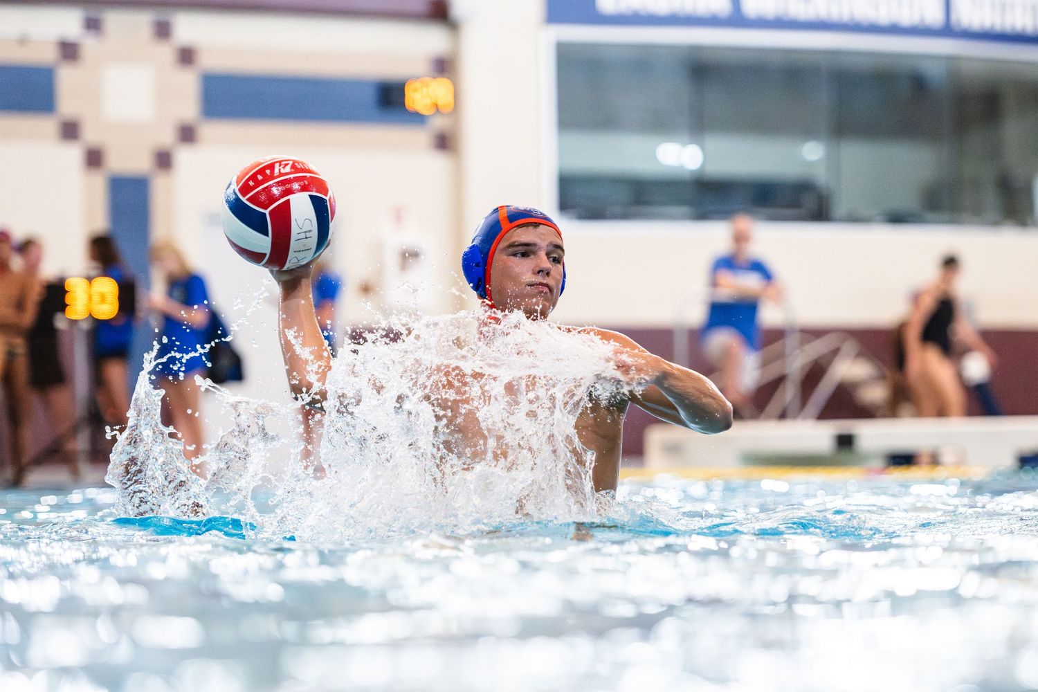 Water polo player winds up to throw ball during splashing competitive match at indoor aquatic championship event.