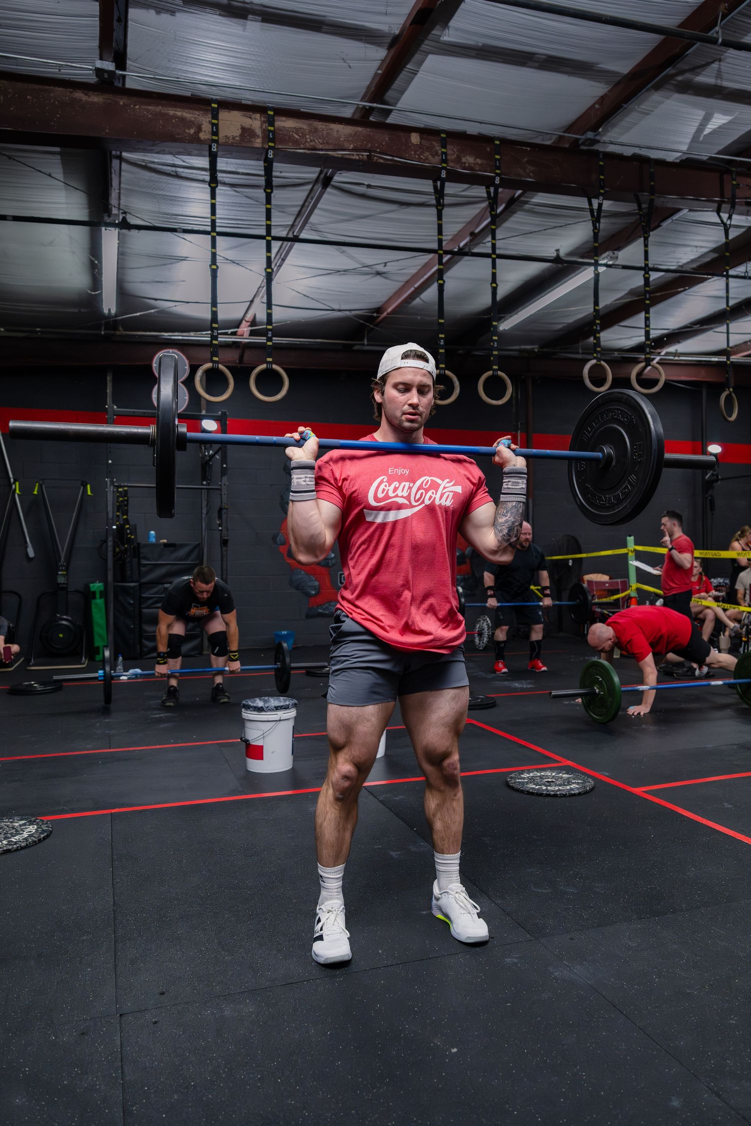 Muscular man in Coca-Cola shirt performing a barbell lift at a CrossFit gym.