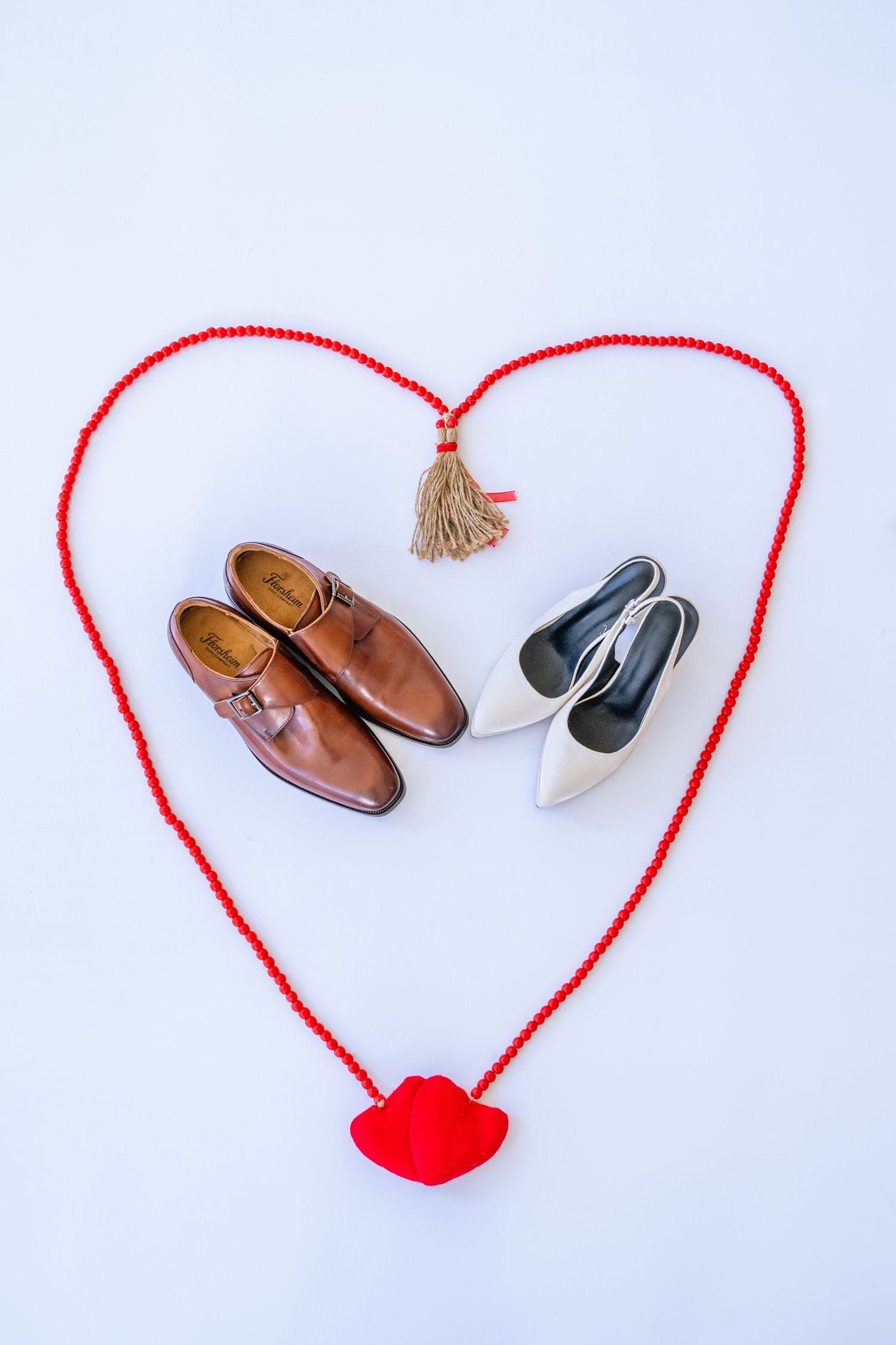 Brown men's dress shoes and white bridal heels arranged inside a red beaded heart shape on white background.