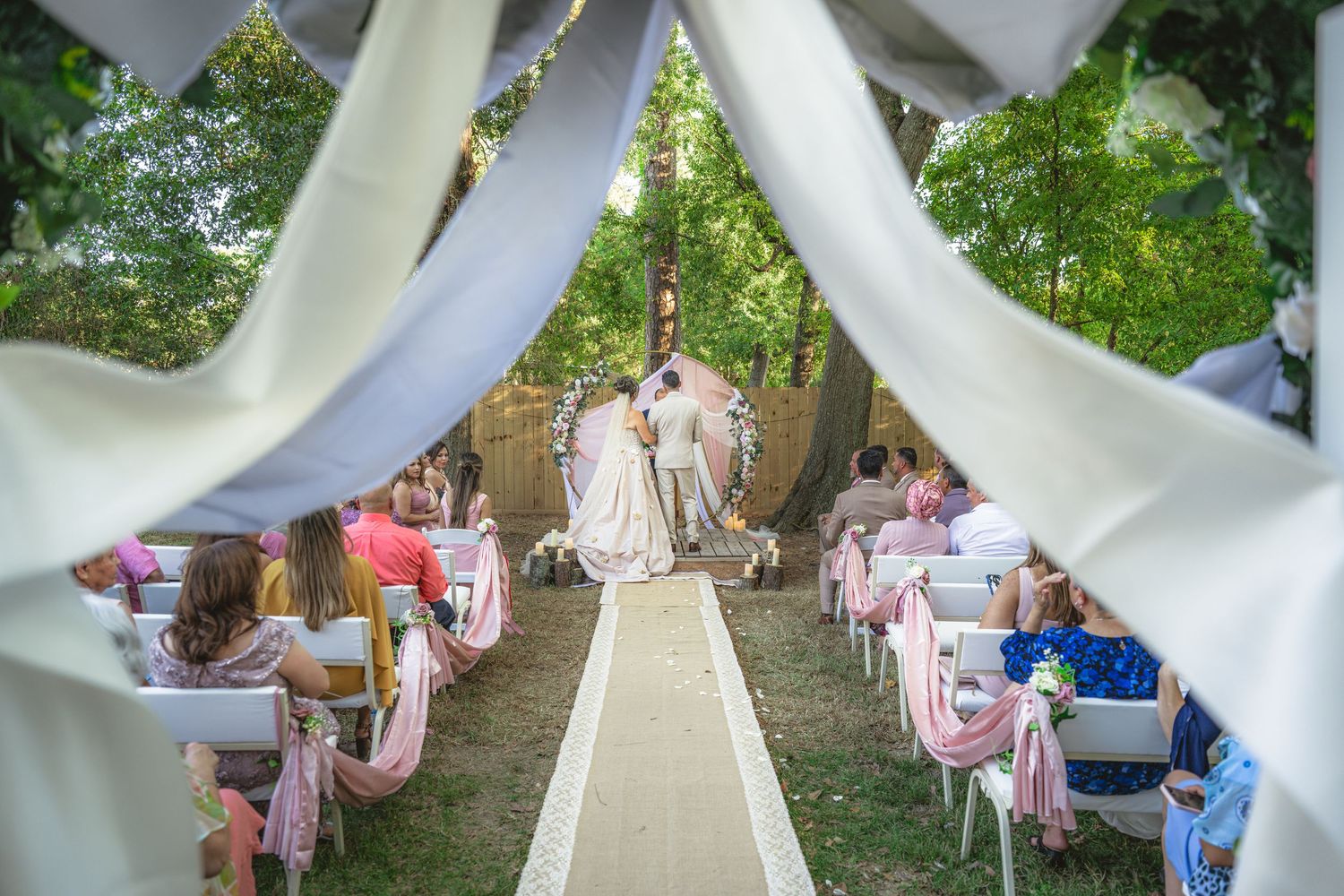 Elegant outdoor wedding ceremony with white draping, pink ribbon chairs, and couple at the altar surrounded by lush green trees.