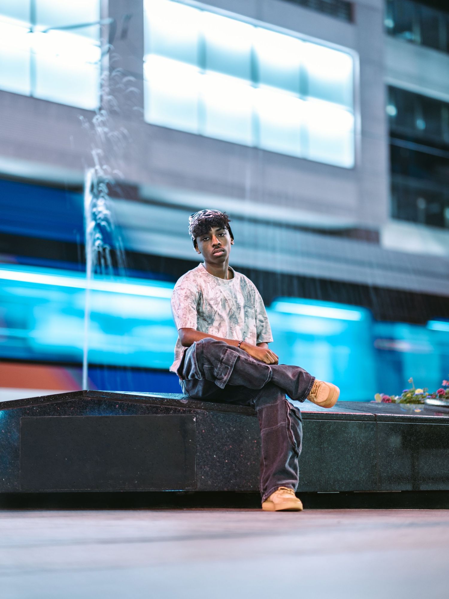 Young man sitting on urban fountain ledge with blurred city lights and rain in background.