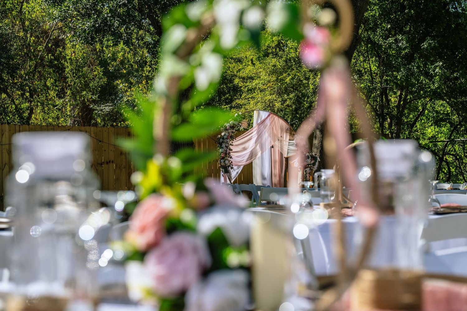 Elegant outdoor wedding reception table with pink floral arrangements and a draped canopy arch amid lush green trees.