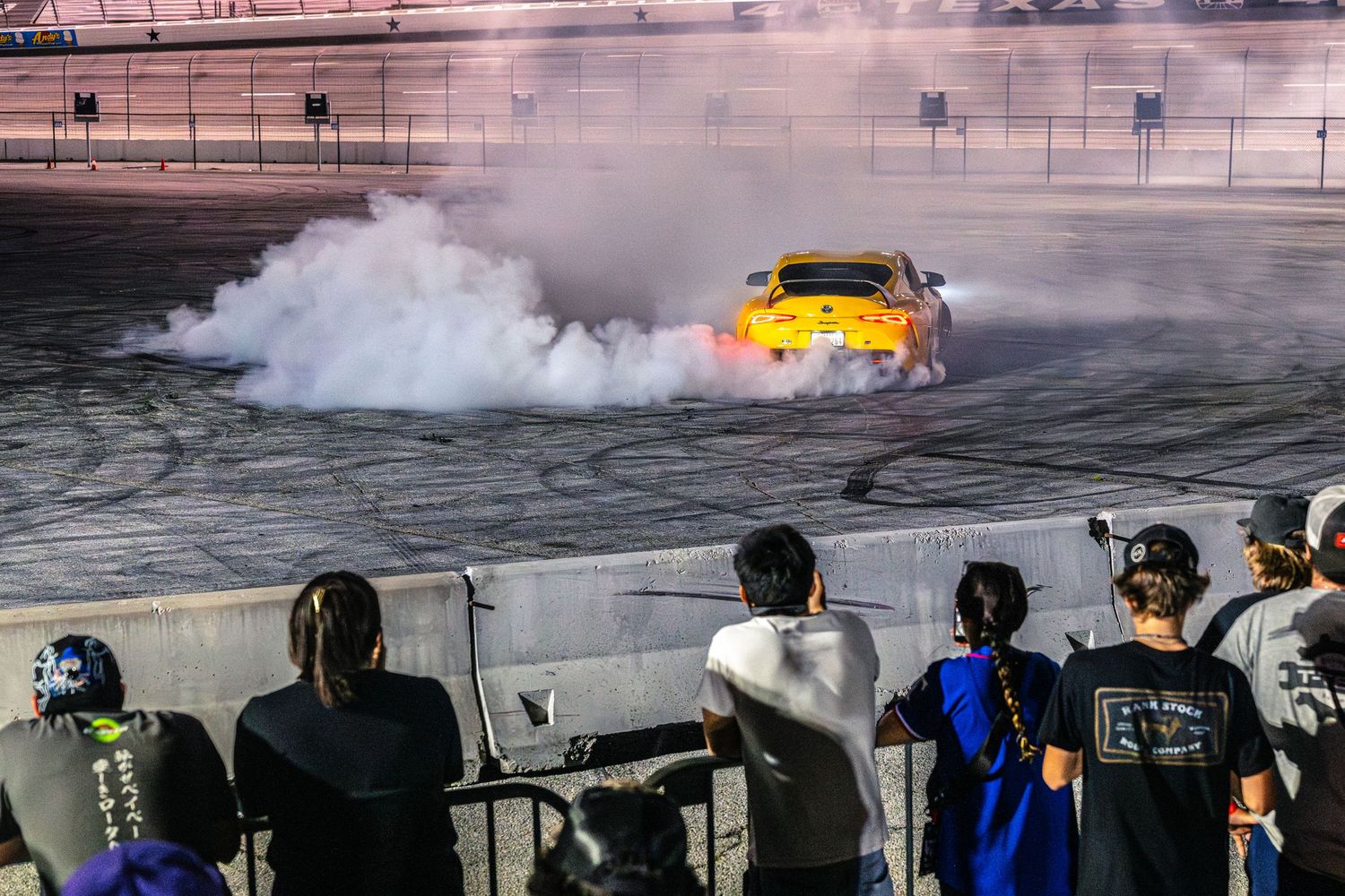 Yellow sports car doing a massive burnout on a racetrack while spectators watch from behind a wall.