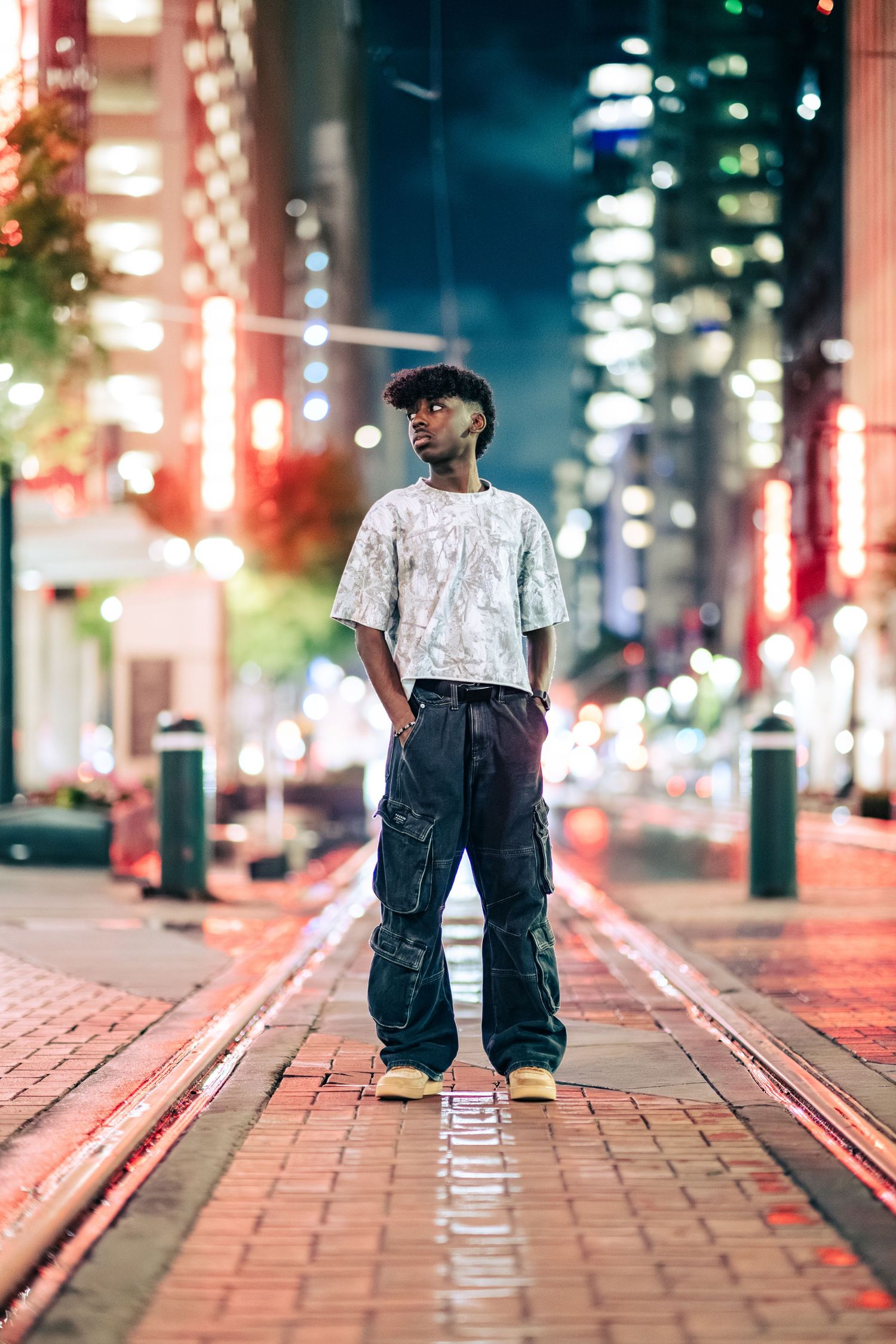 Young man in cargo pants standing on illuminated city street at night with skyline.