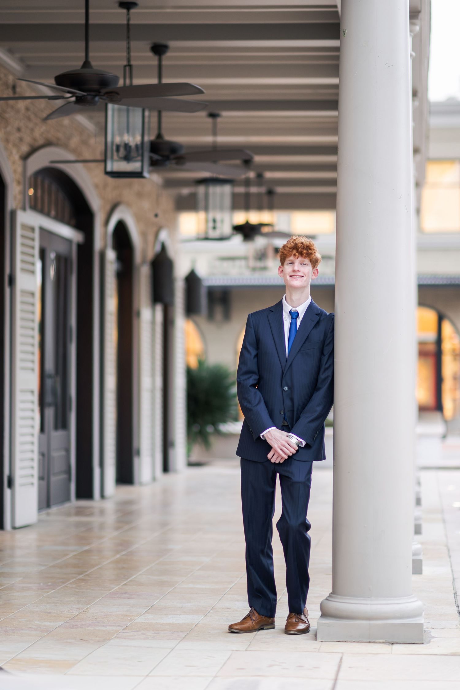 Smiling redhead teen in navy suit stands by column in elegant outdoor corridor.
