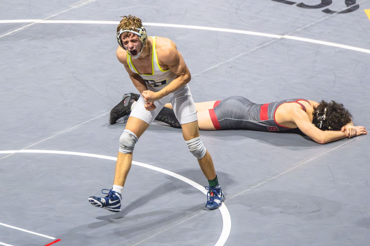 Two wrestlers compete on a mat, one standing triumphantly while opponent lies flat during a wrestling match.