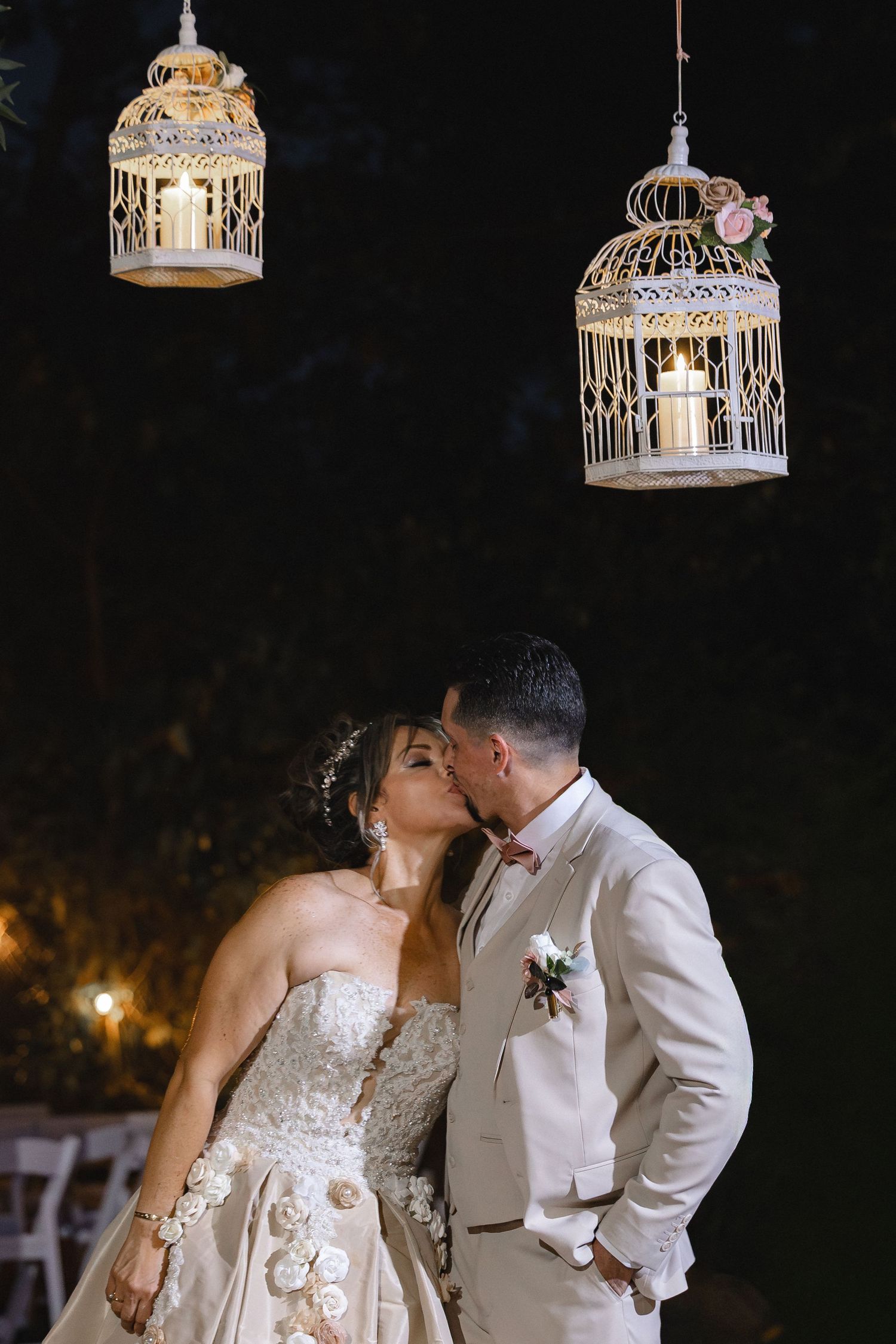 Bride and groom share a romantic kiss under glowing decorative bird cage lanterns at an elegant outdoor night wedding.