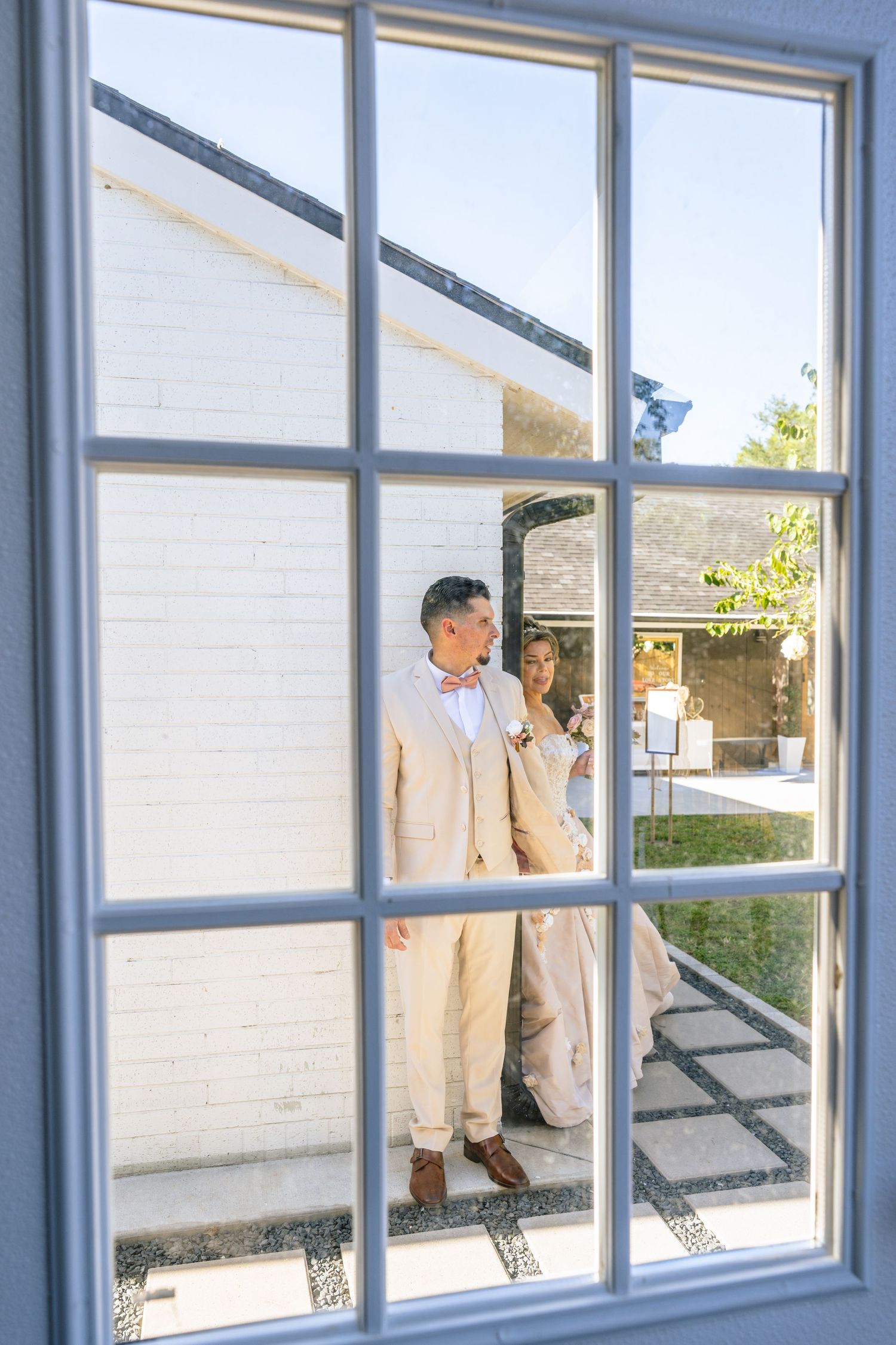 Groom in beige suit and pink bow tie seen through white-framed window at outdoor wedding venue.