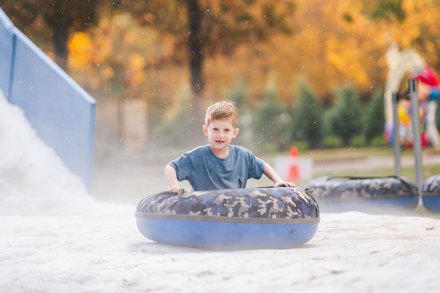 Boy smiling while riding a blue inflatable tube down a snow slide into sandy landing area outdoors.