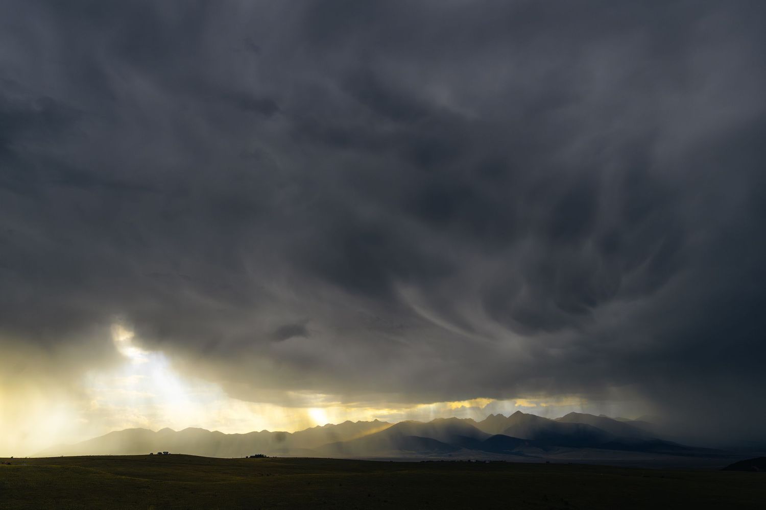Ominous dark storm clouds with golden light rays over a silhouetted mountain range at dusk.