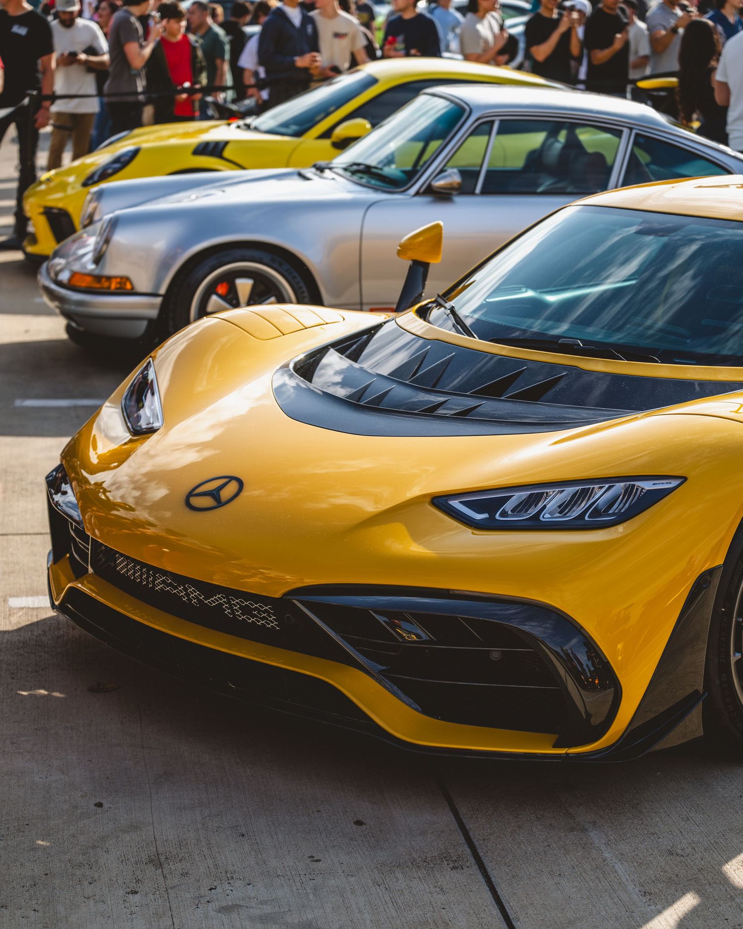 Yellow Mercedes-AMG ONE hypercar at an exotic car show with a silver Porsche 911 visible behind it.