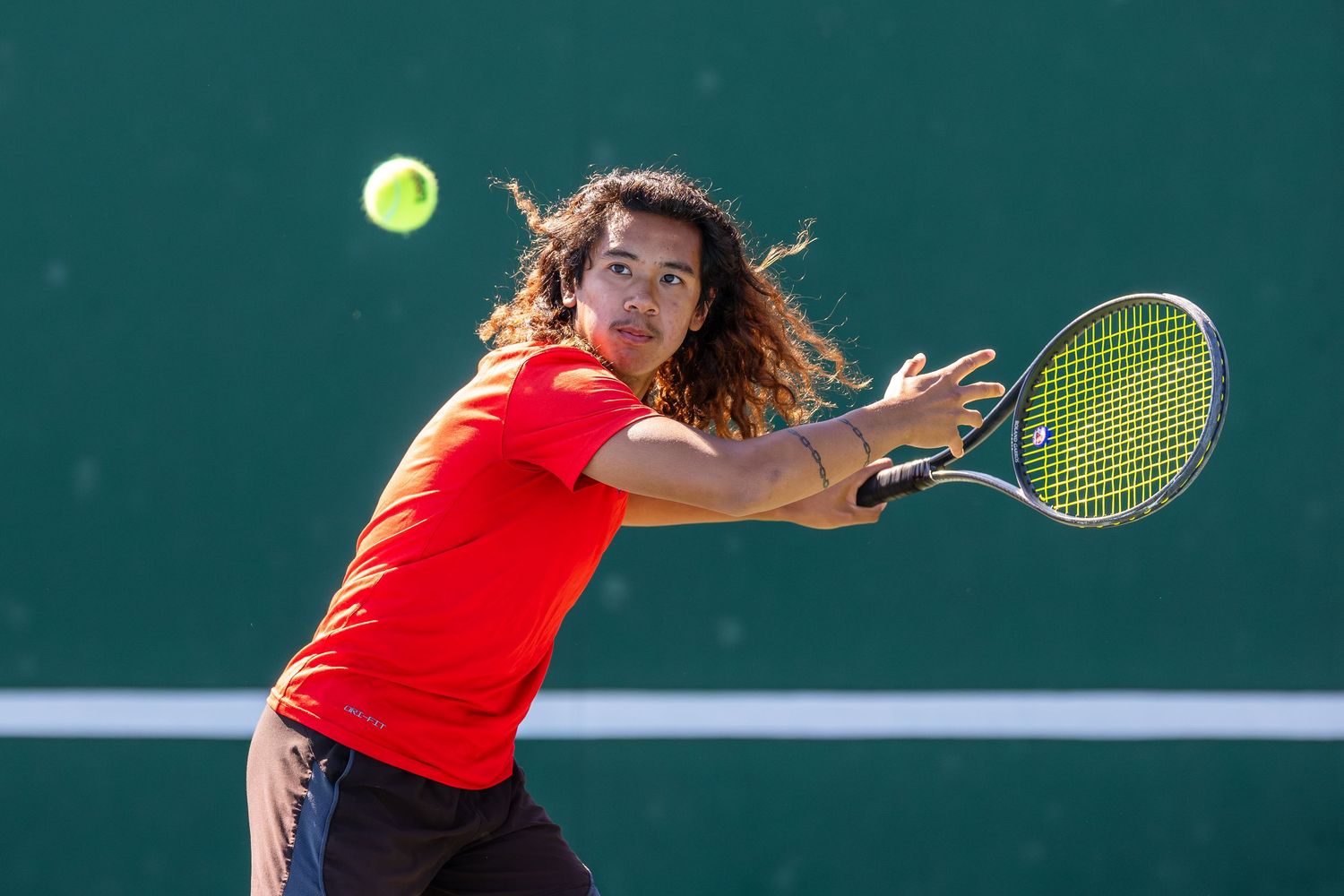 Young tennis player with curly hair in red shirt hits a backhand shot on an outdoor court.