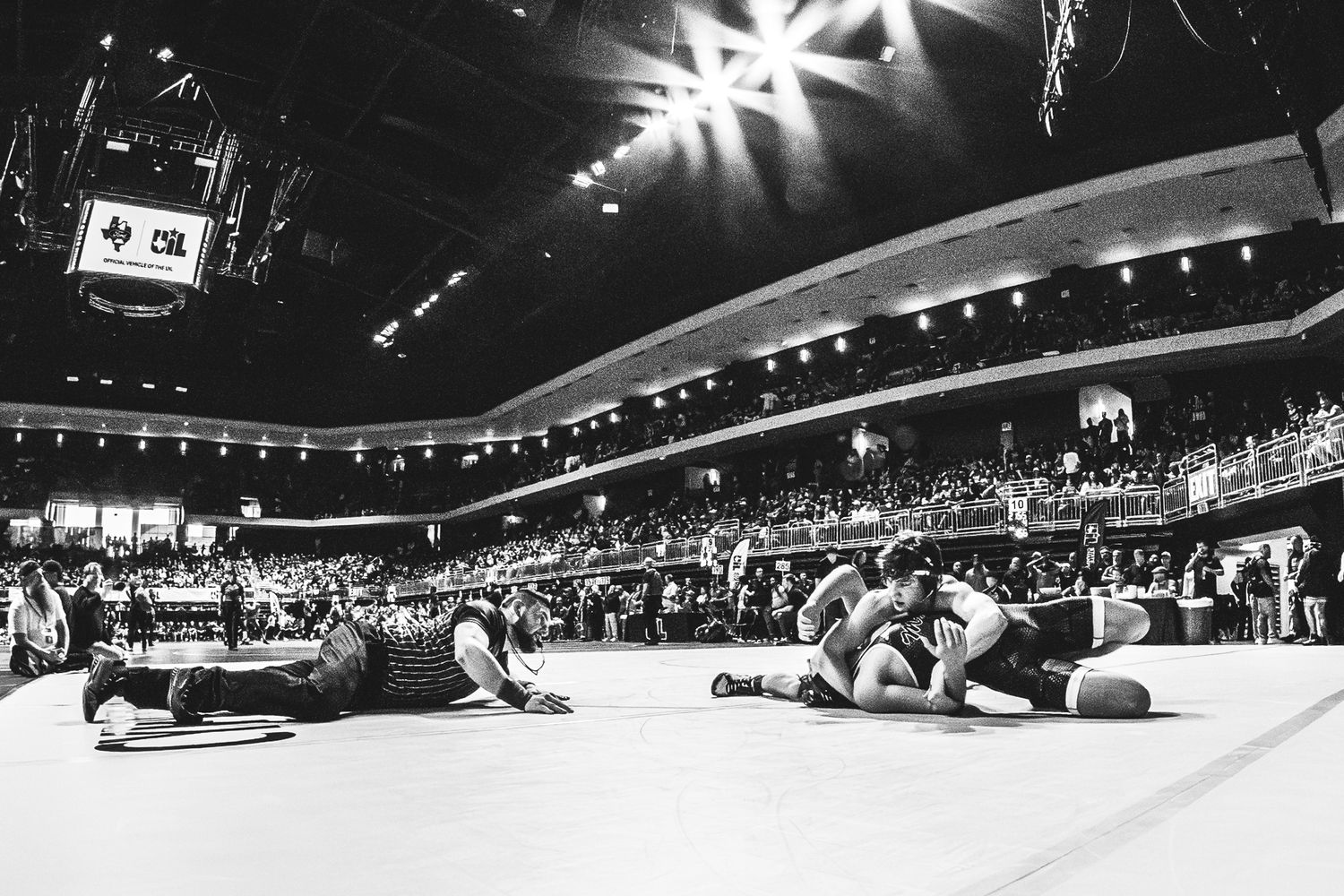 Black and white photo of wrestlers competing on a mat in a large arena filled with spectators.