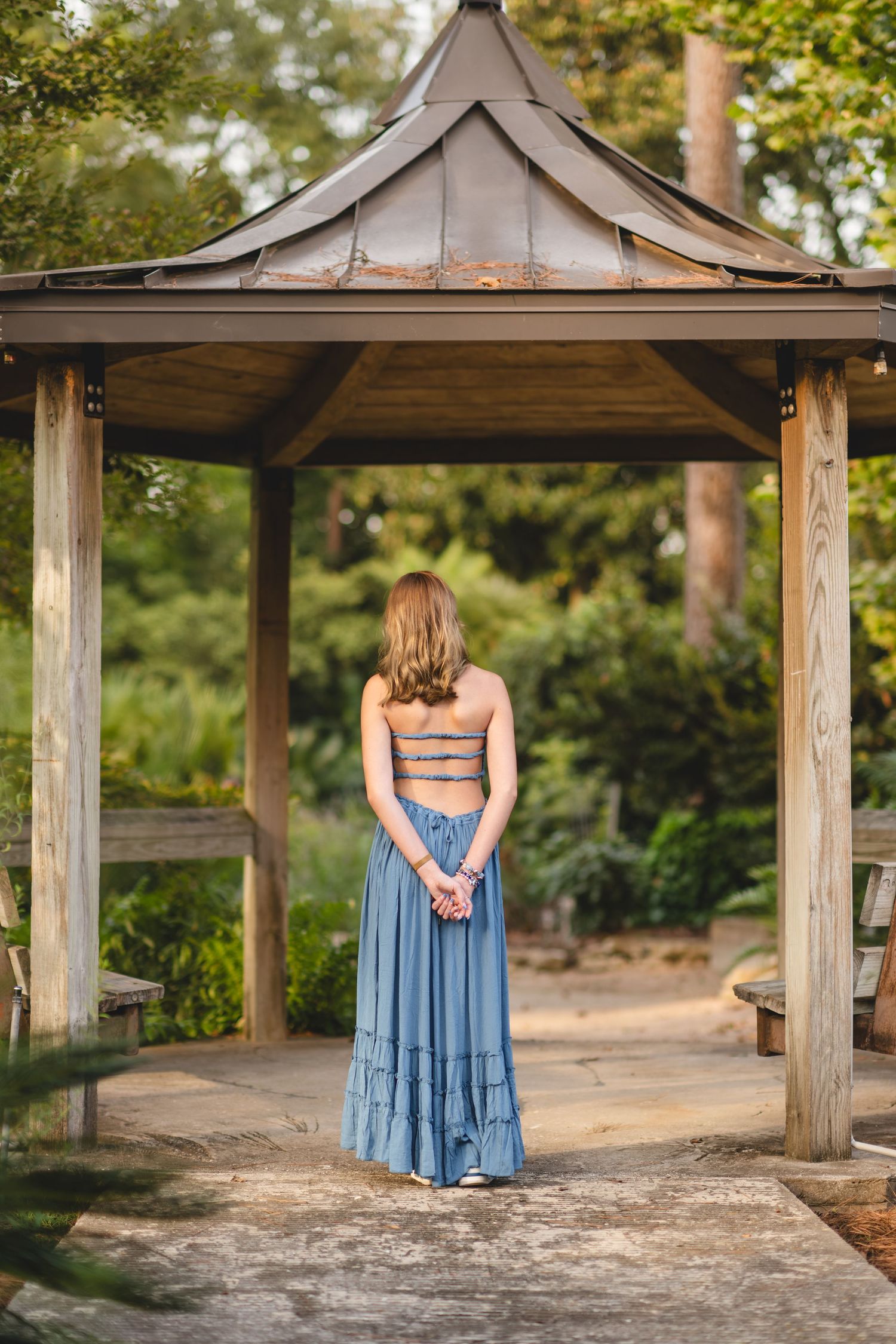 Young woman in blue maxi skirt stands with back turned under wooden garden gazebo.