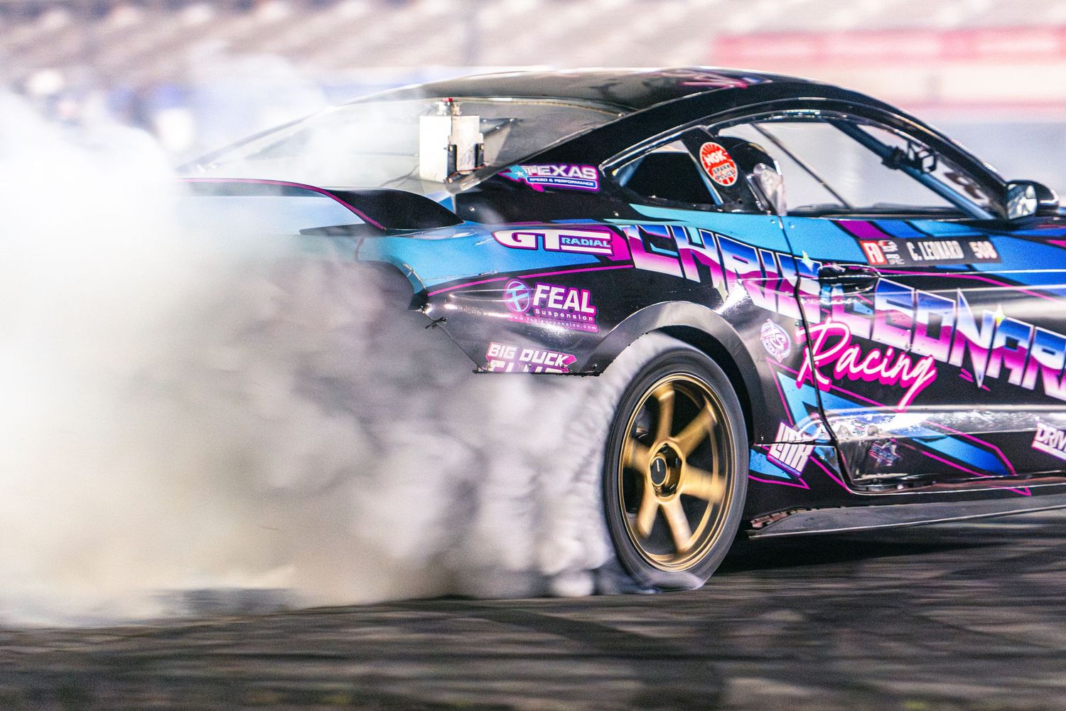 A drift car with purple and blue racing livery creates massive tire smoke on a motorsport track.