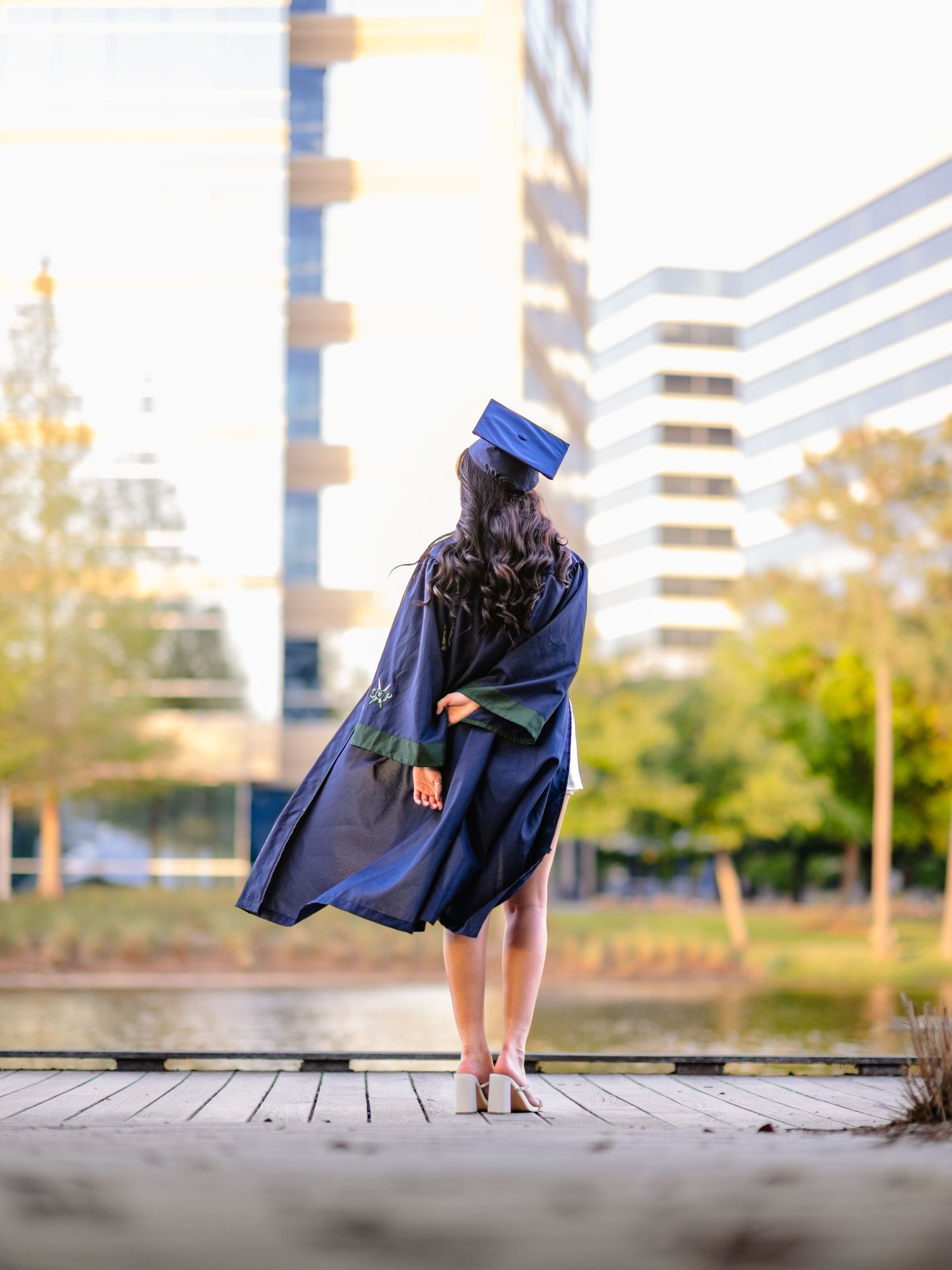 Graduate in navy cap and gown standing on wooden dock with city buildings in background.