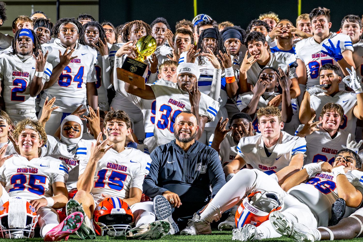 Football team and coach pose together celebrating a victory on the field after winning a game.