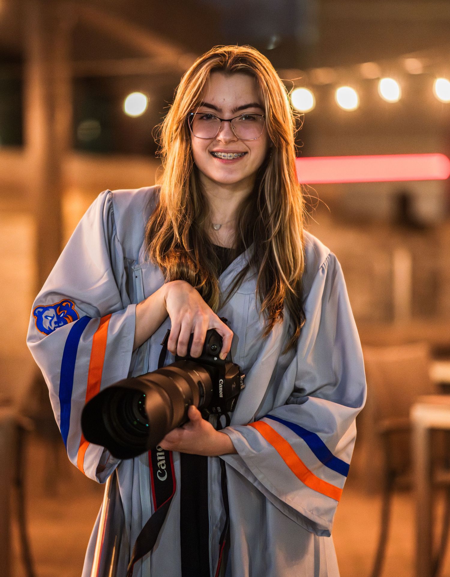 Smiling teen in graduation gown holding a Canon camera with bokeh lights in the background.
