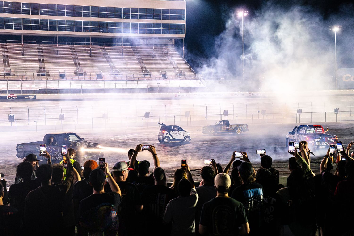 Multiple cars performing simultaneous burnouts at a nighttime motorsport event as crowd watches and films.