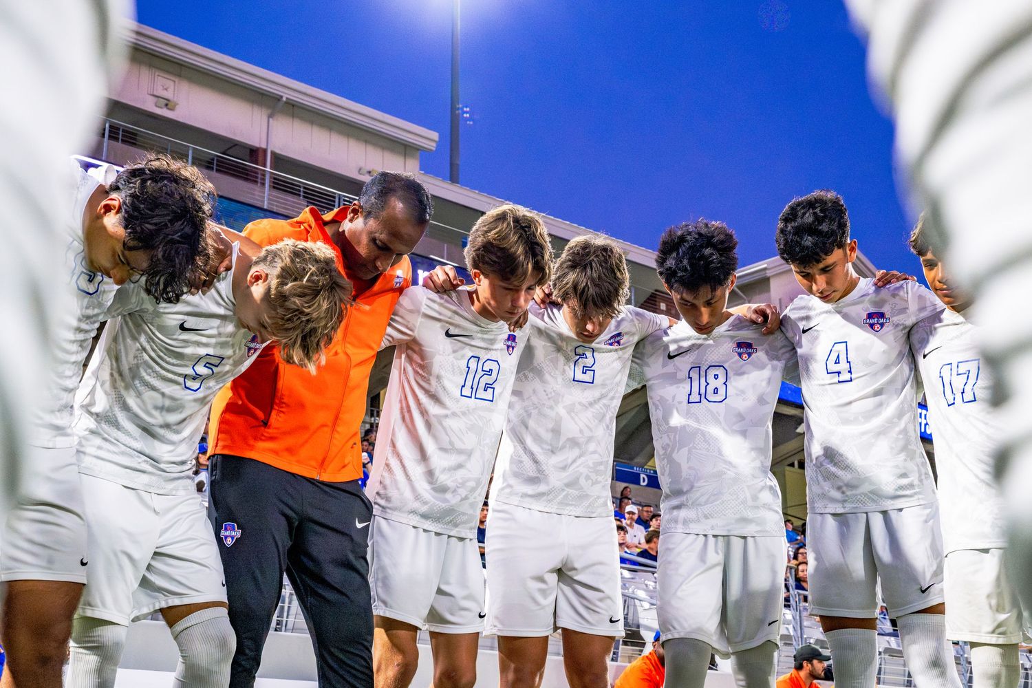 Boys soccer team huddles together with coach under blue stadium lights after a match.