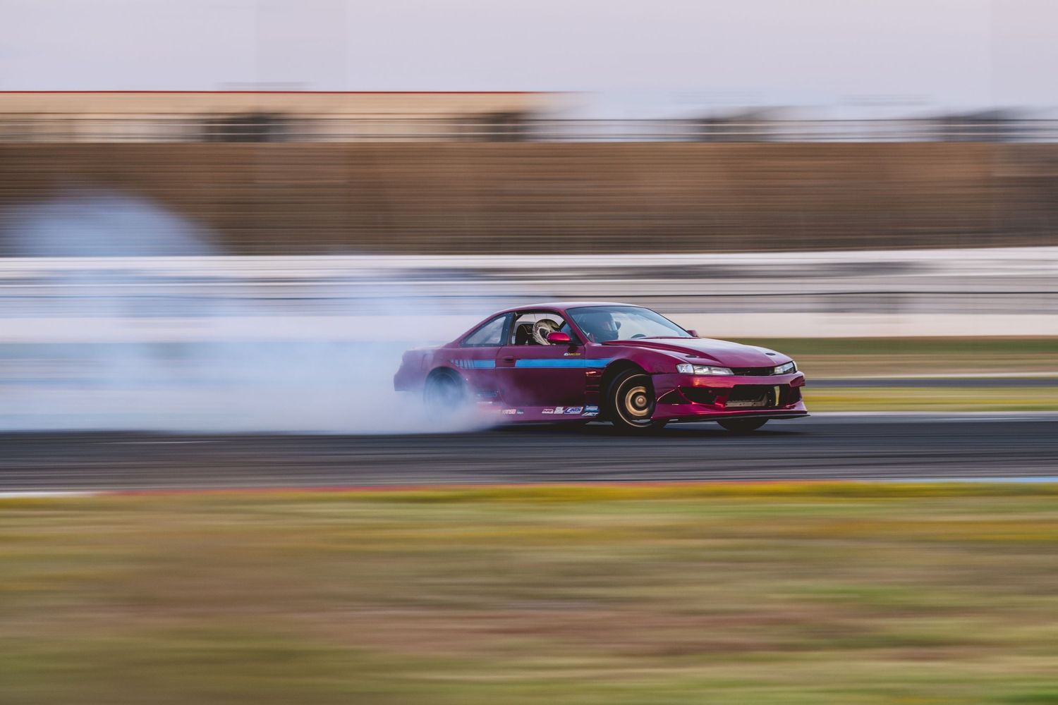 A dark red car produces thick tire smoke while drifting on a racetrack during daytime motorsport event.