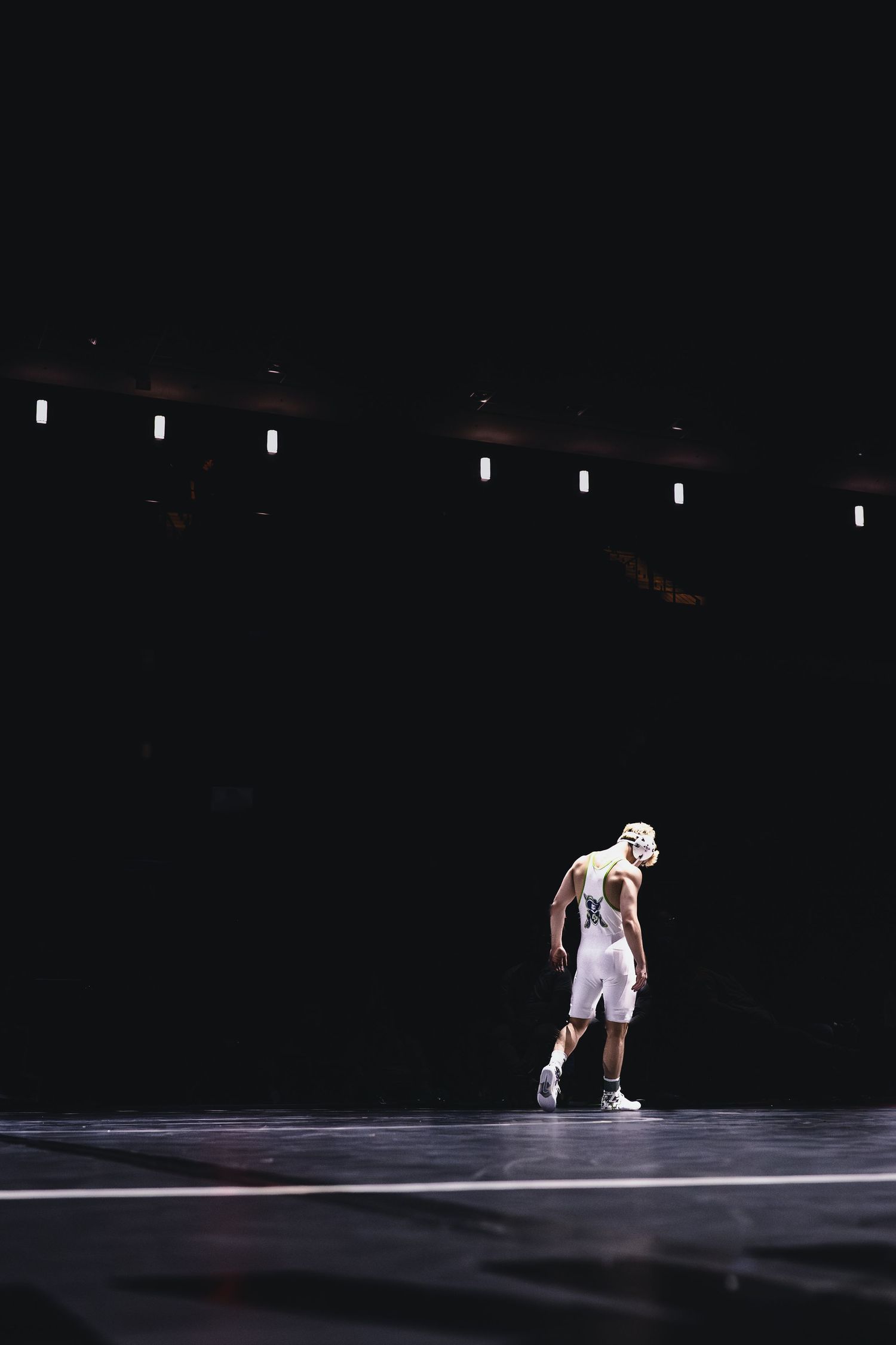 Wrestler in white singlet and headgear stands alone on dark arena court.