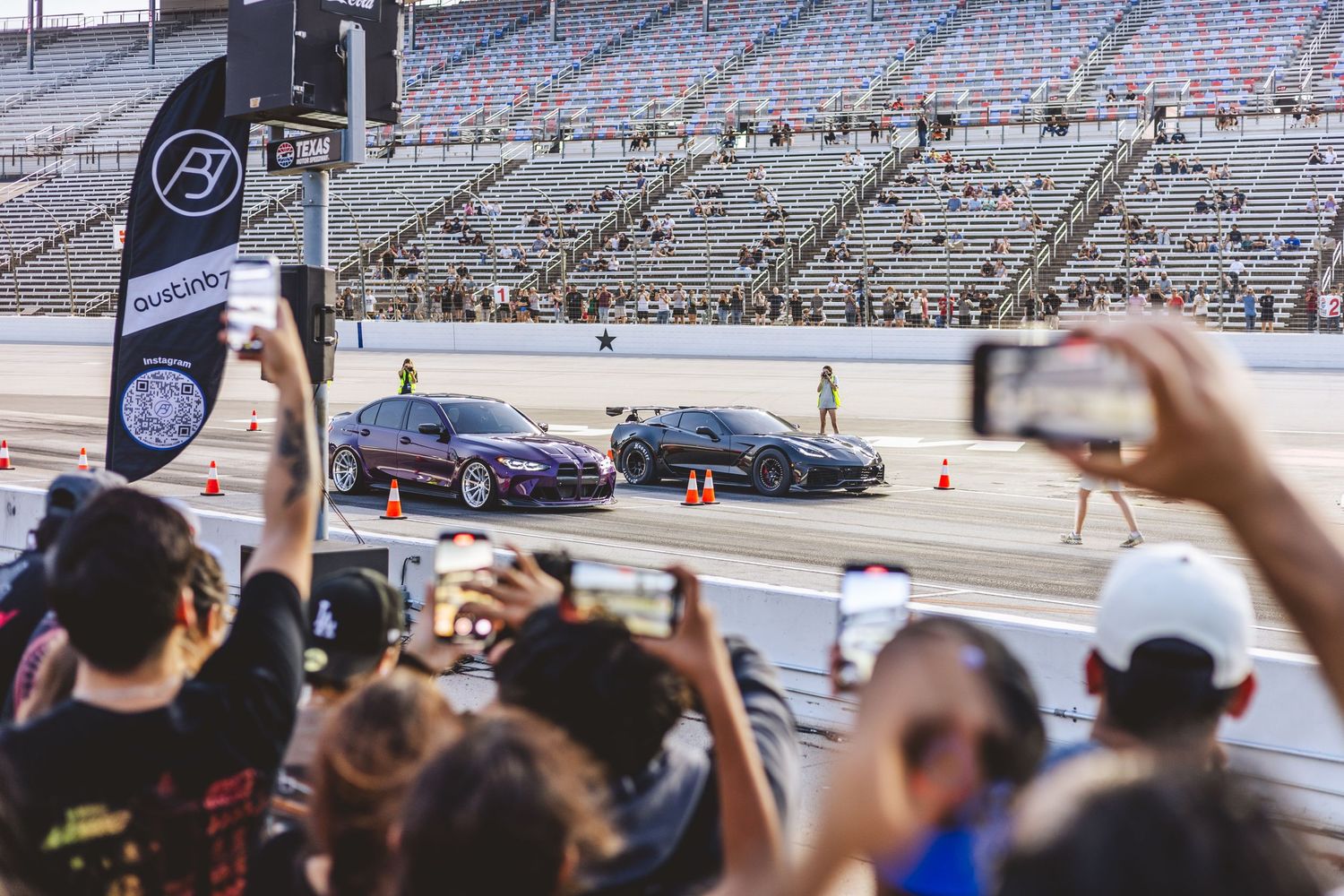 Spectators photograph two sports cars lined up at the starting line of a drag race at a large stadium.